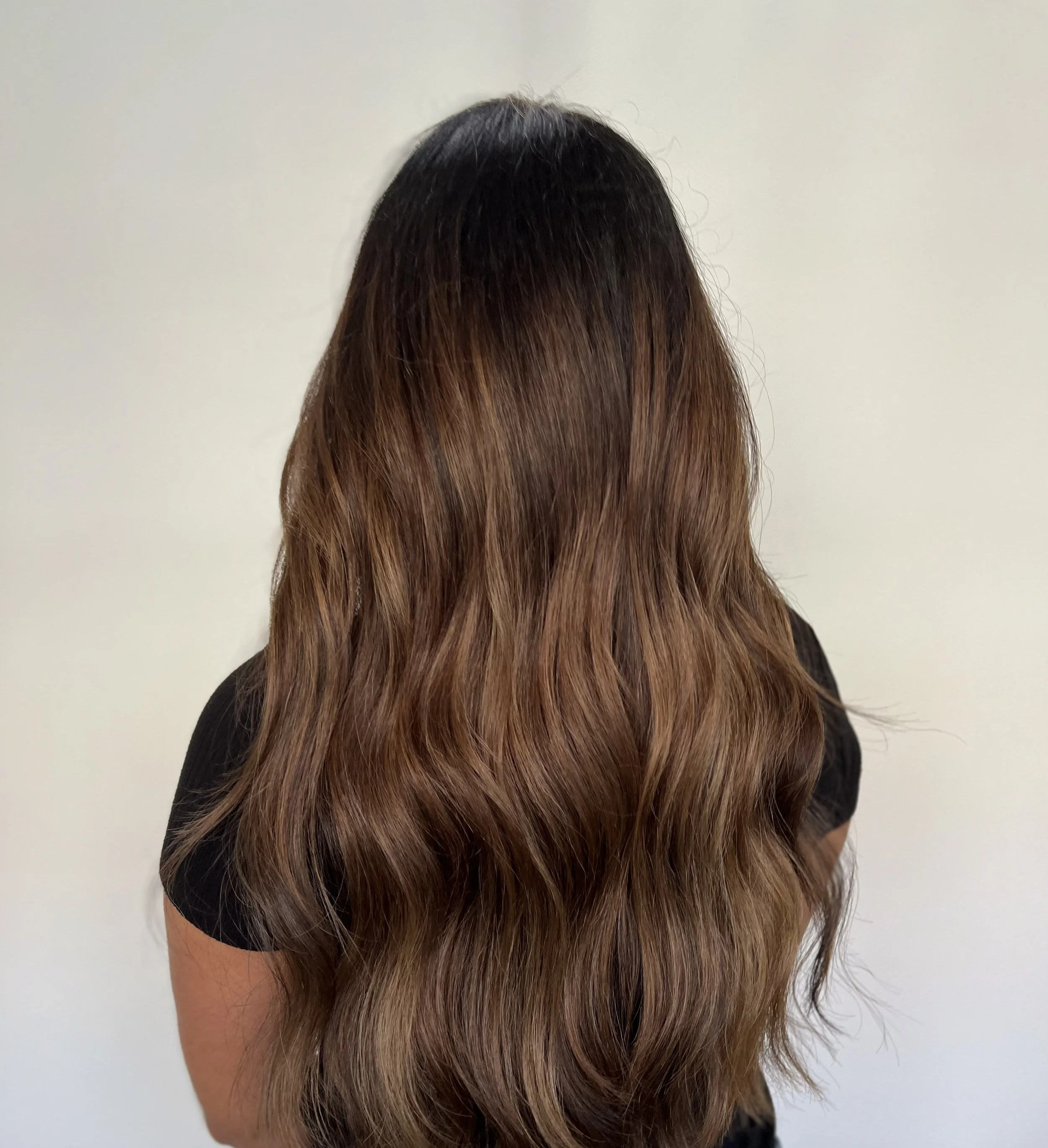 Back of a woman with long, wavy hair that is dark at the roots and gradually lightens to a caramel brown at the ends.