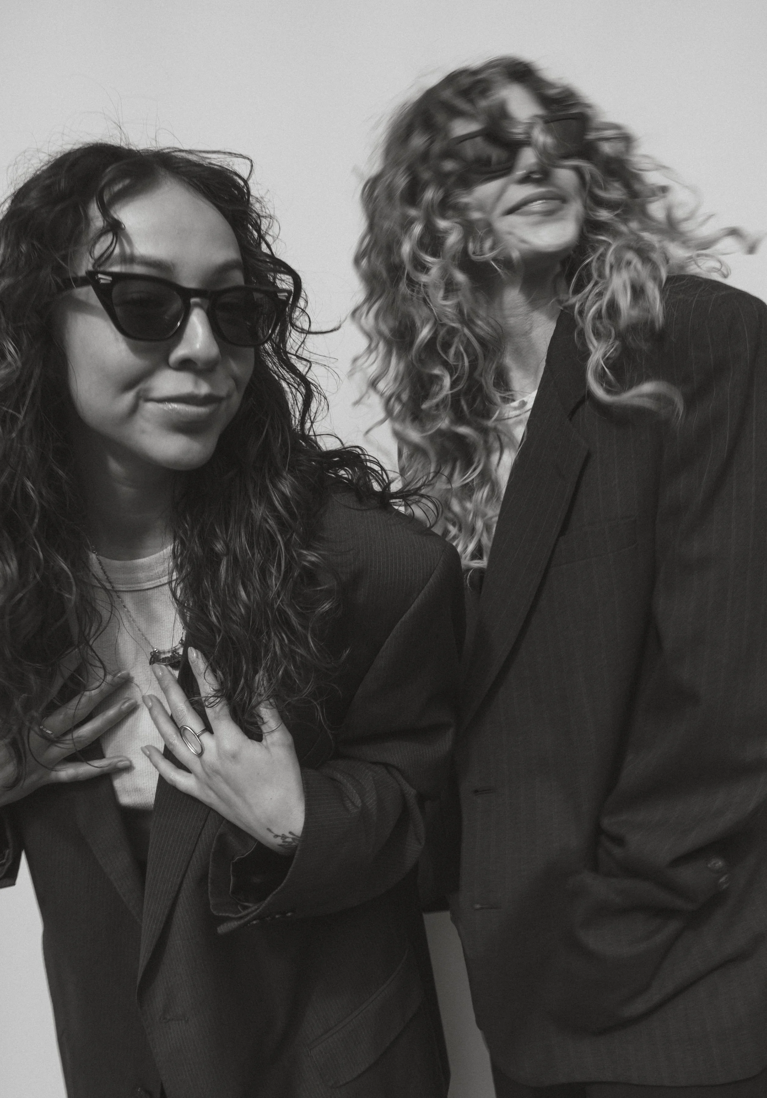Two women with curly hair and sunglasses wearing suits, posing together in black and white.