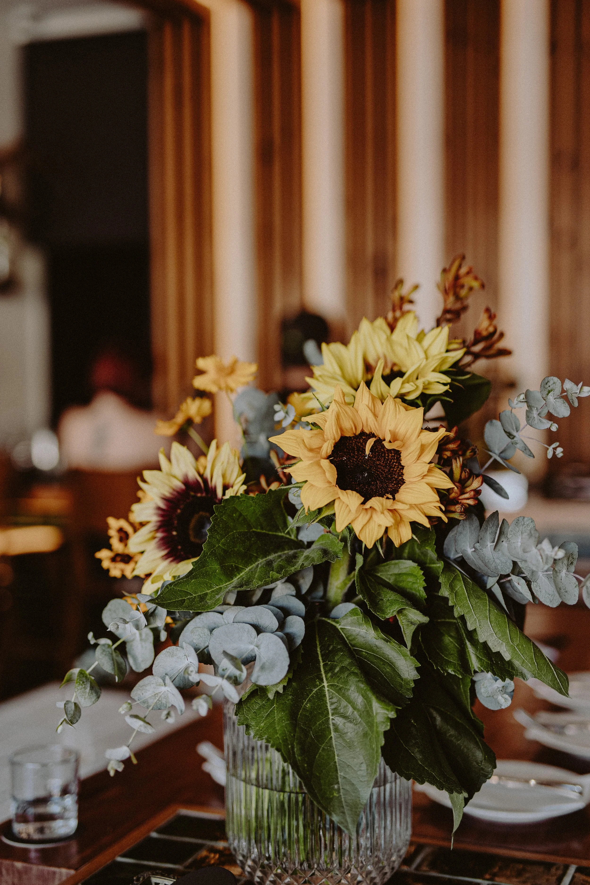 A floral centerpiece with sunflowers, daisies, eucalyptus, and large green leaves on a wooden table.