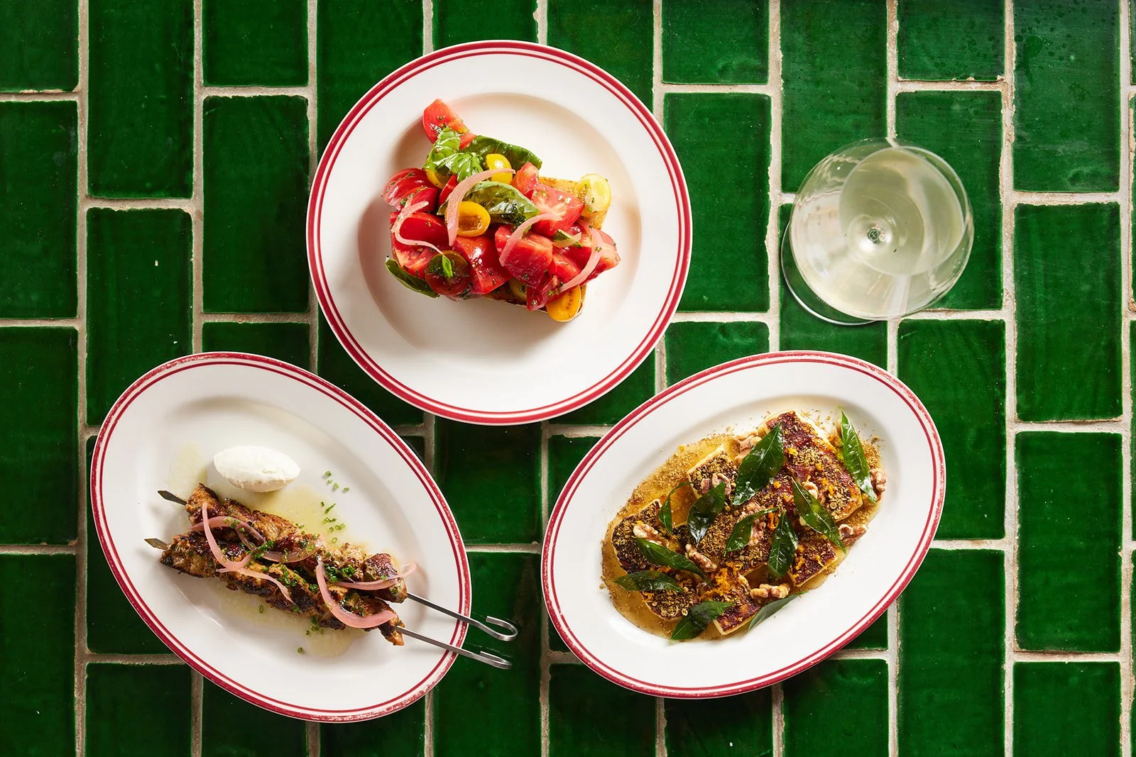 Three plates of food, a glass of white wine, on a green tiled table. The plates have various dishes including salad, grilled meat, and cooked fish garnished with herbs.