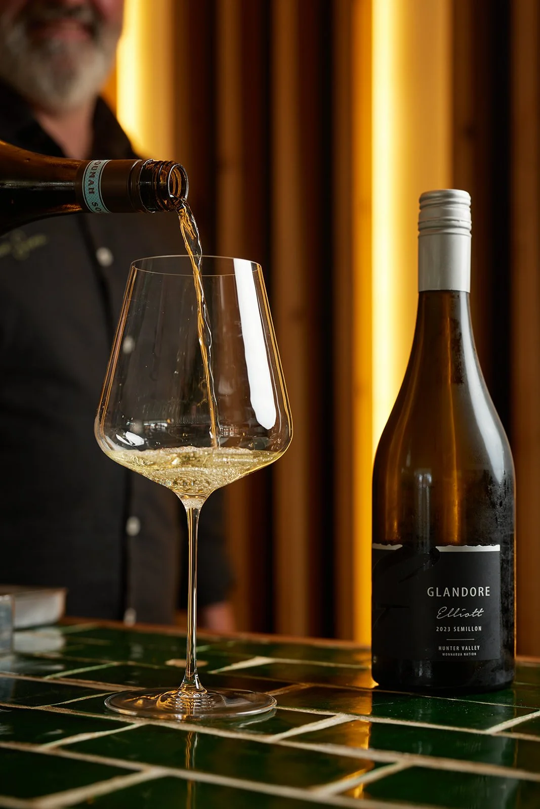 A person pouring white wine from a bottle into a wine glass on a green tiled table, with a bottle of Glandore Elliott wine on the table.
