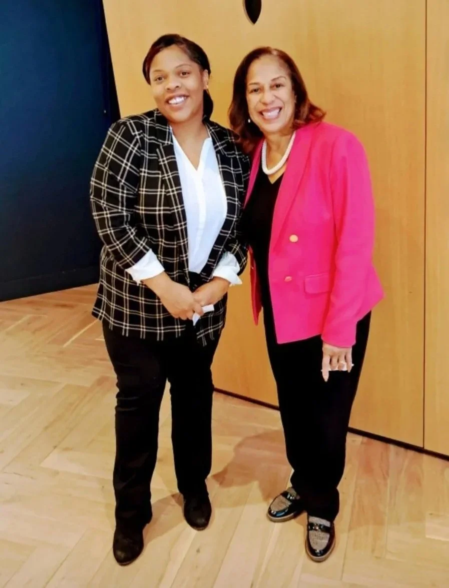 Angel and colleage standing together indoors, smiling at the camera. One wears a black and white checked blazer with white shirt and black pants. The other wears a bright pink blazer with black pants and a white necklace.