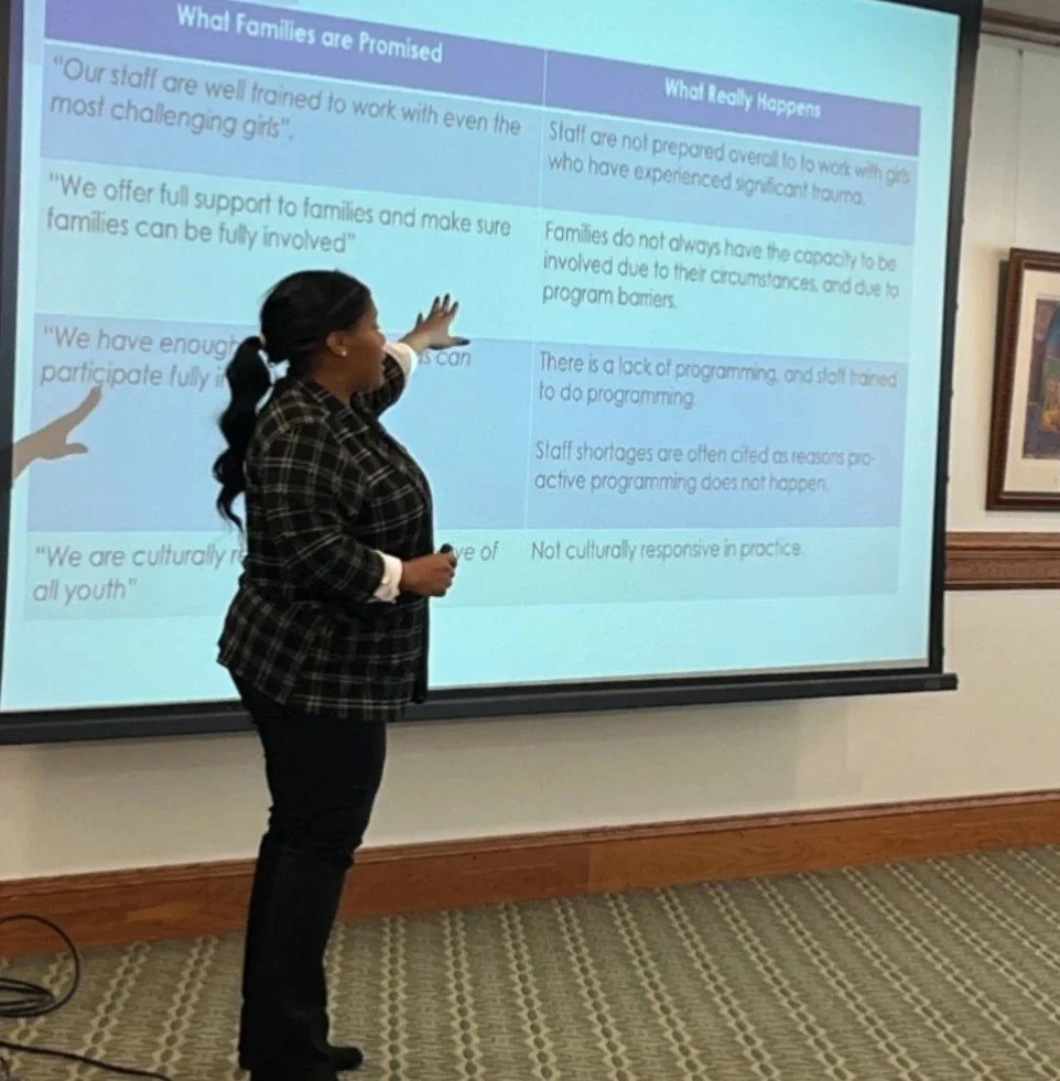 A woman in a black and white plaid jacket standing in front of a projector screen, pointing at a presentation slide. The slide compares promises made to families with what actually happens, including issues like trauma and lack of program responsiveness.