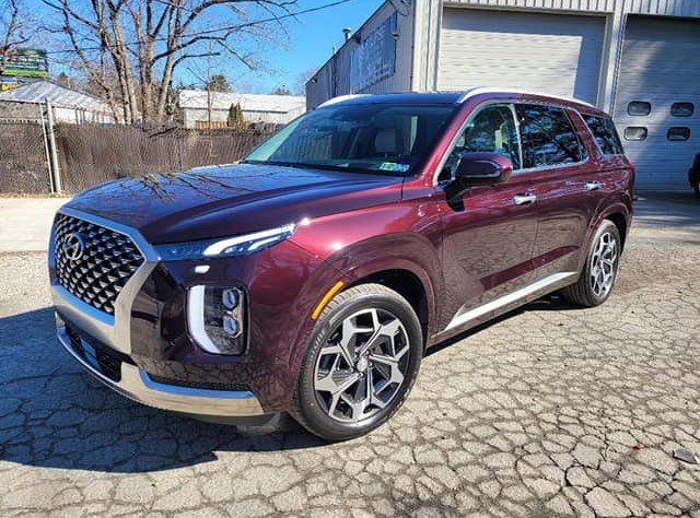 A maroon-colored Hyundai SUV parked on a cracked asphalt driveway in front of a building with a garage door and a fence, with leafless trees in the background.