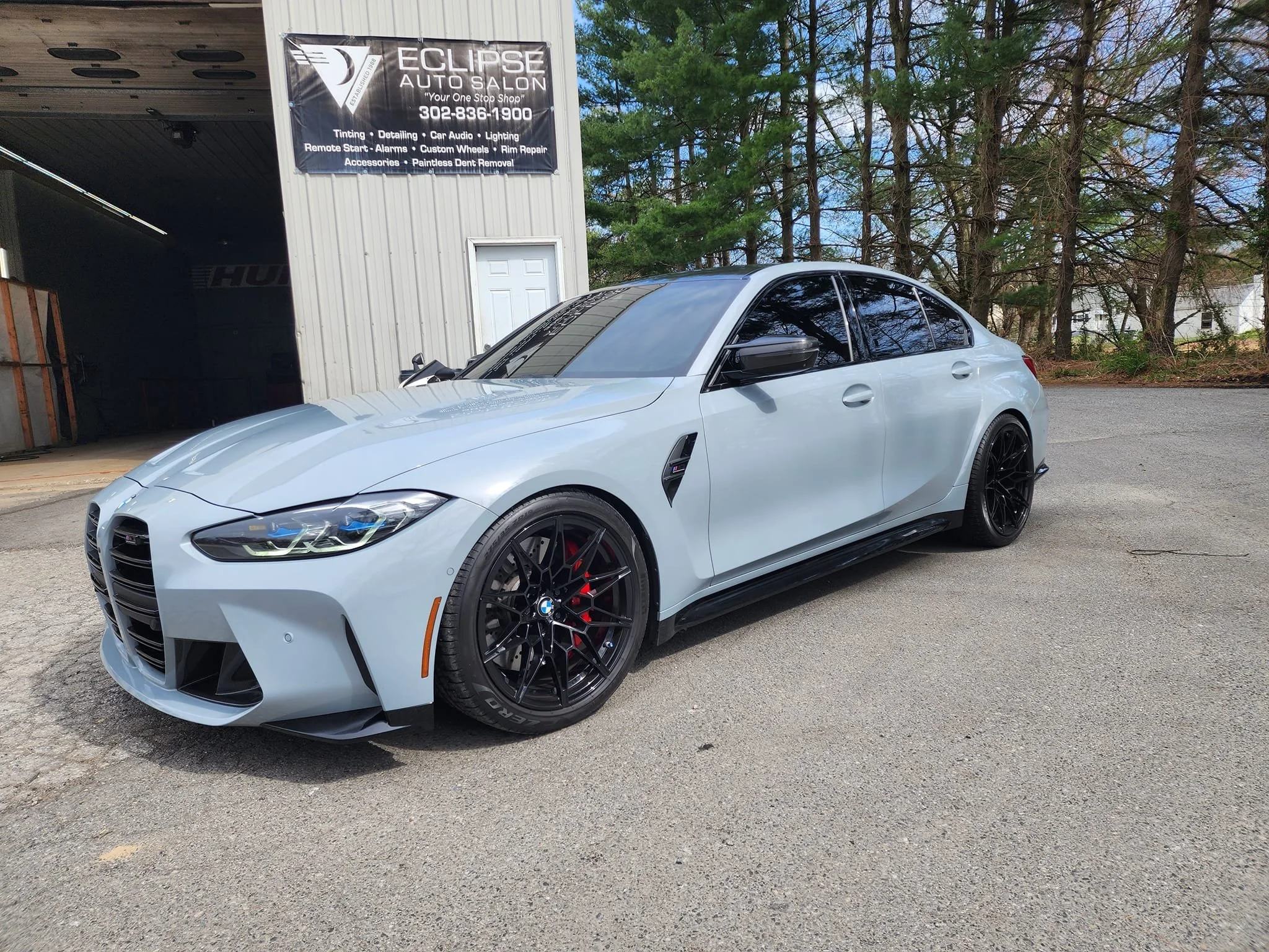 A silver BMW M3 parked outside a garage with a sign reading 'Eclipse Auto Salon' that offers auto detailing, tinting, lighting, and other services.