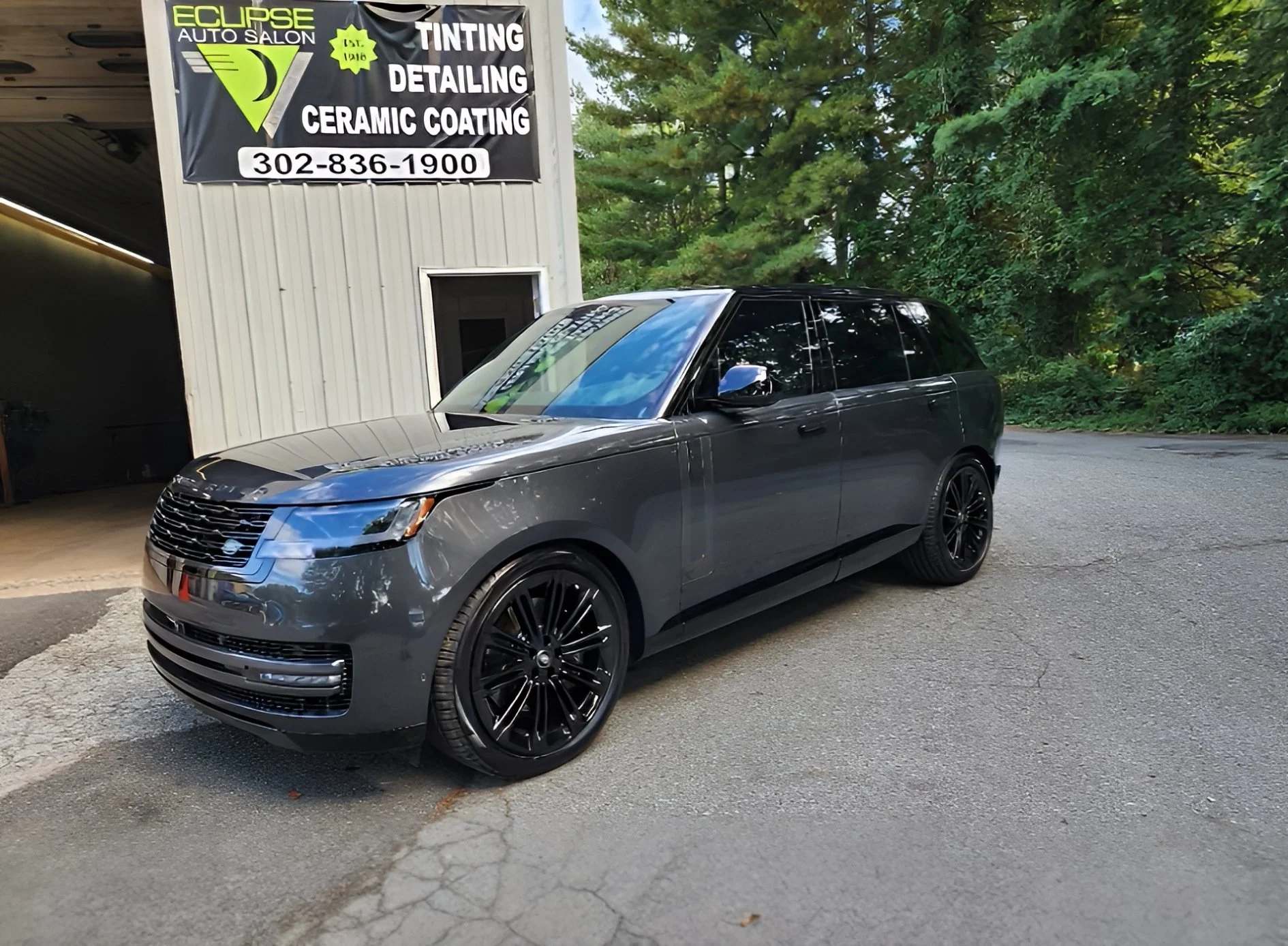 Gray luxury SUV parked outdoors next to a white building with a sign advertising auto services like tinting, detailing, and ceramic coating. There are trees and greenery in the background.