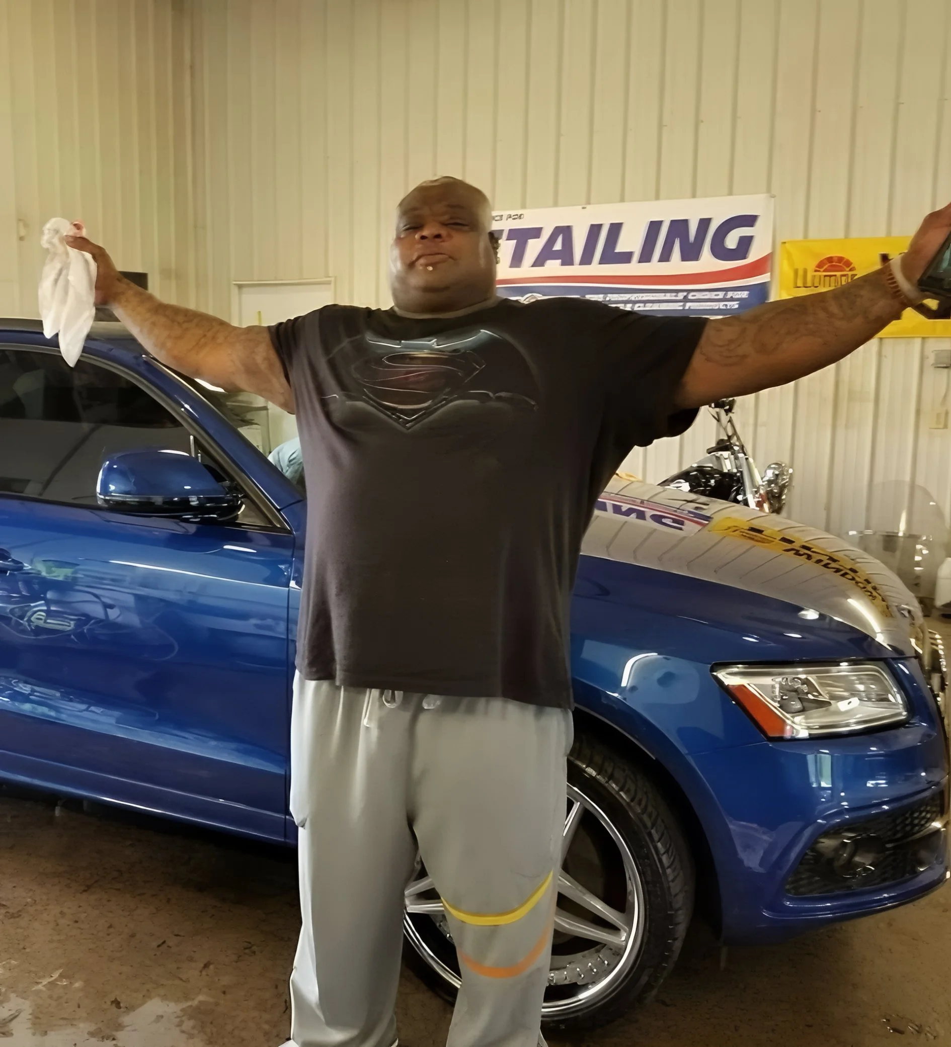 Man with arms outstretched standing in front of a blue car inside a garage with a banner that says 'Detailing' in the background.