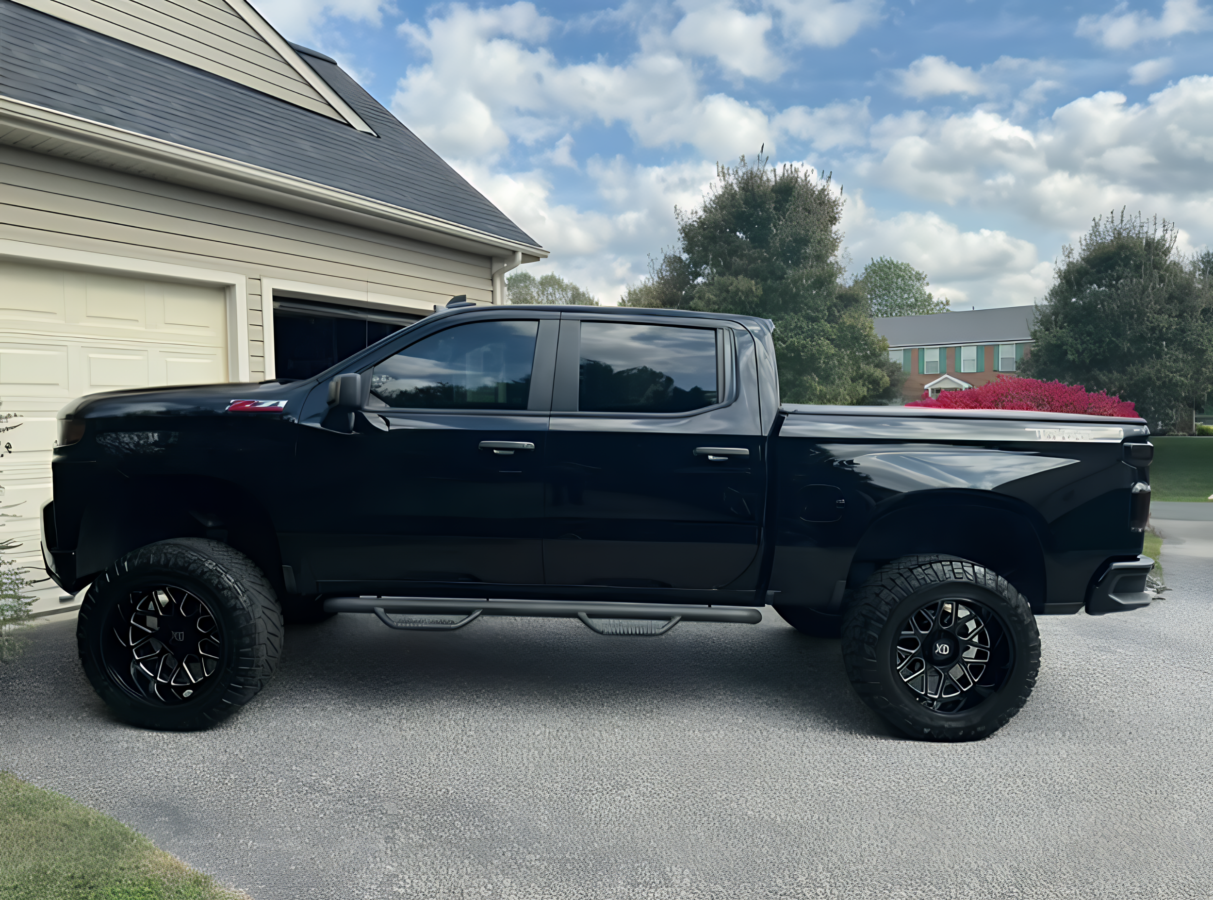 Black pickup truck parked in a driveway next to a house with a garage, under a partly cloudy sky.