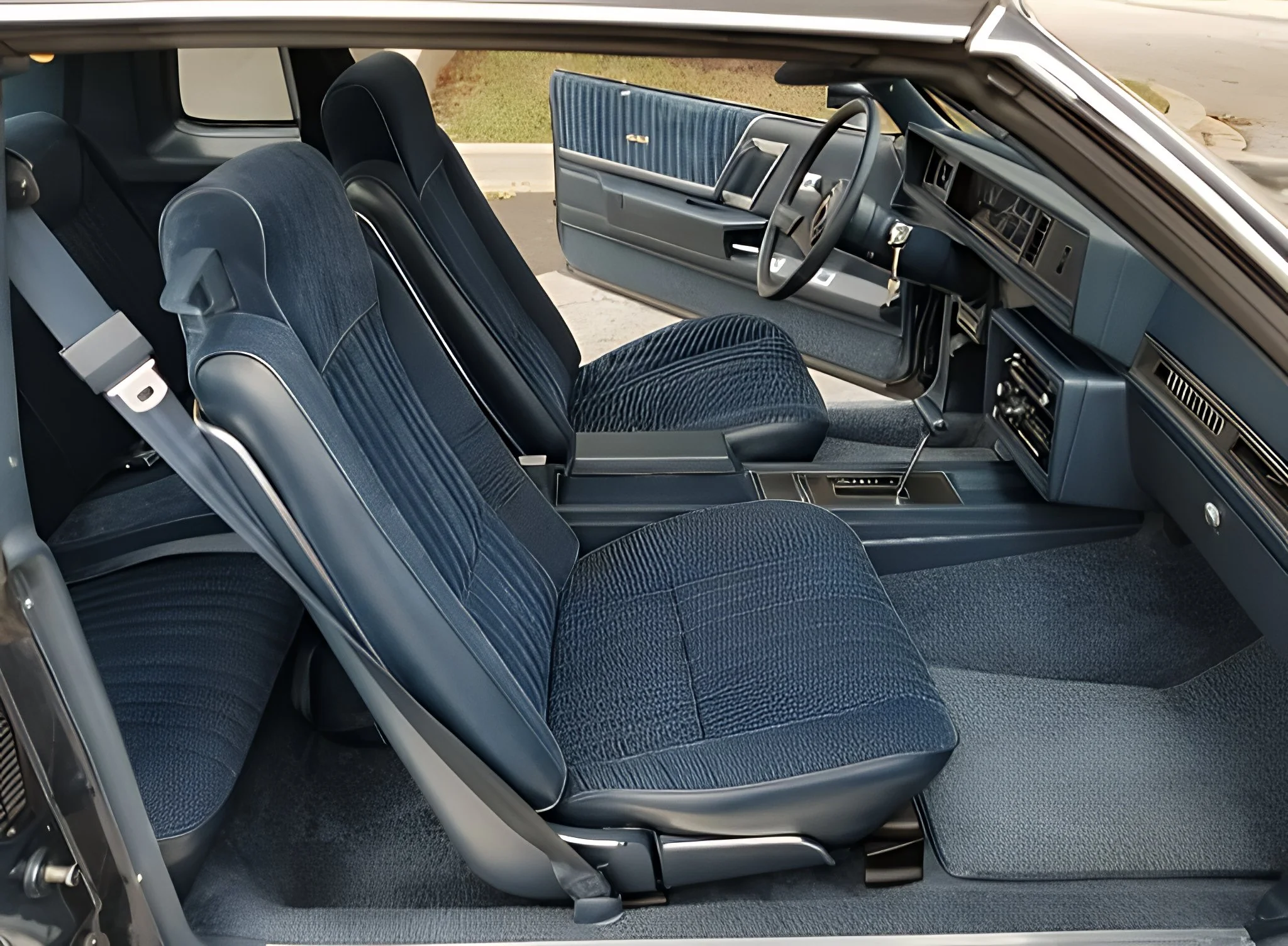 Interior of a vintage car showing front seats, dashboard, steering wheel, and open driver-side door with blue and black upholstery.