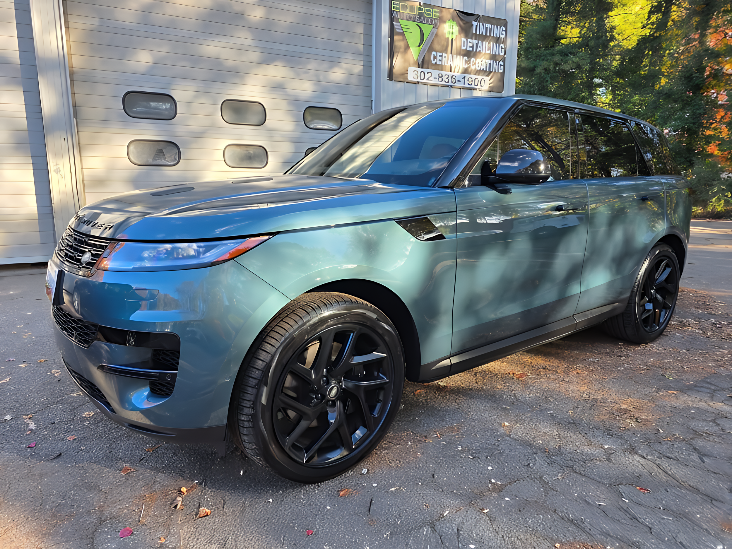 A modern blue Range Rover SUV parked on pavement outside a garage with a sign for auto tinting, detailing, and ceramic coating.