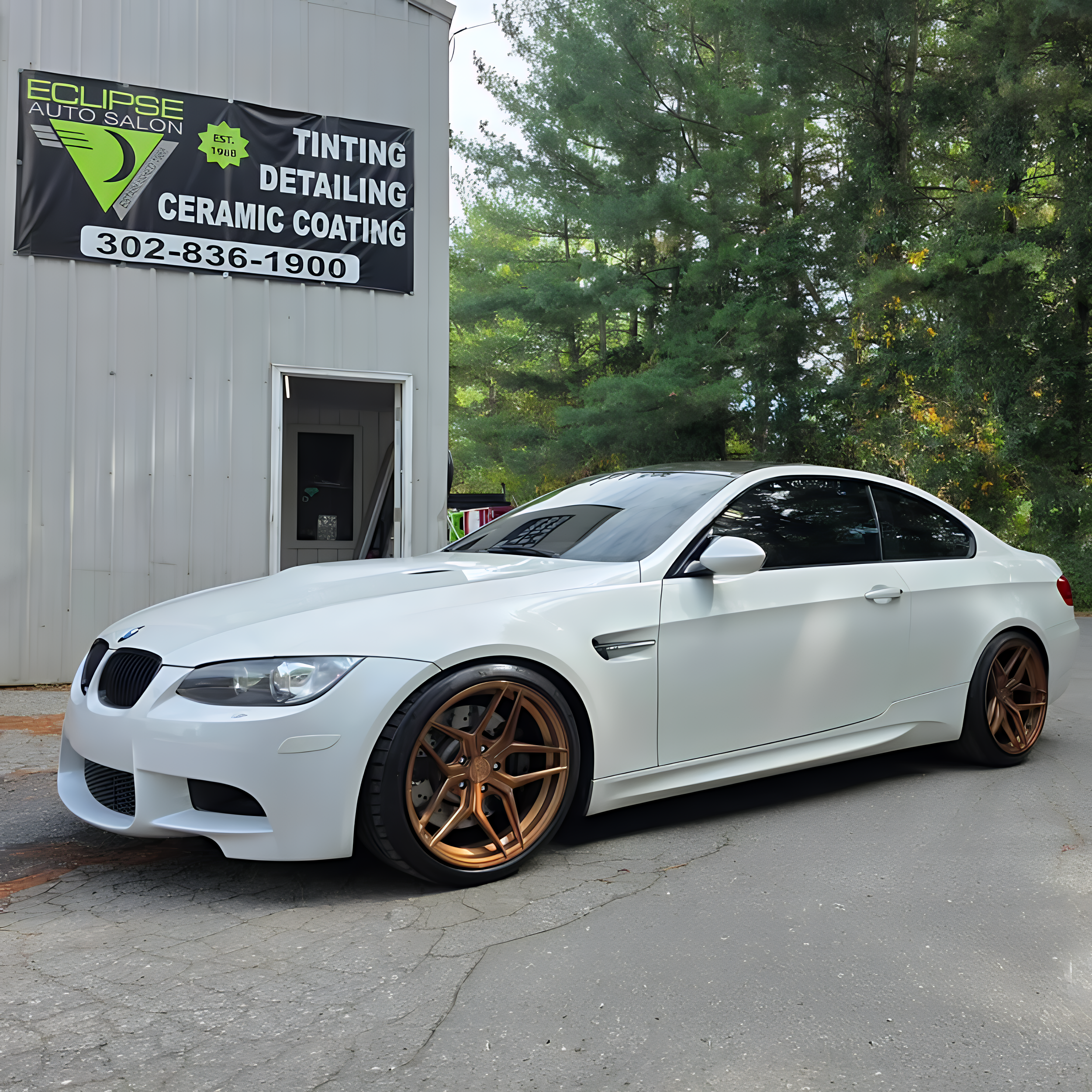 A white sports car with bronze-colored wheels parked in front of an auto salon with a sign advertising tinting, detailing, and ceramic coating services.