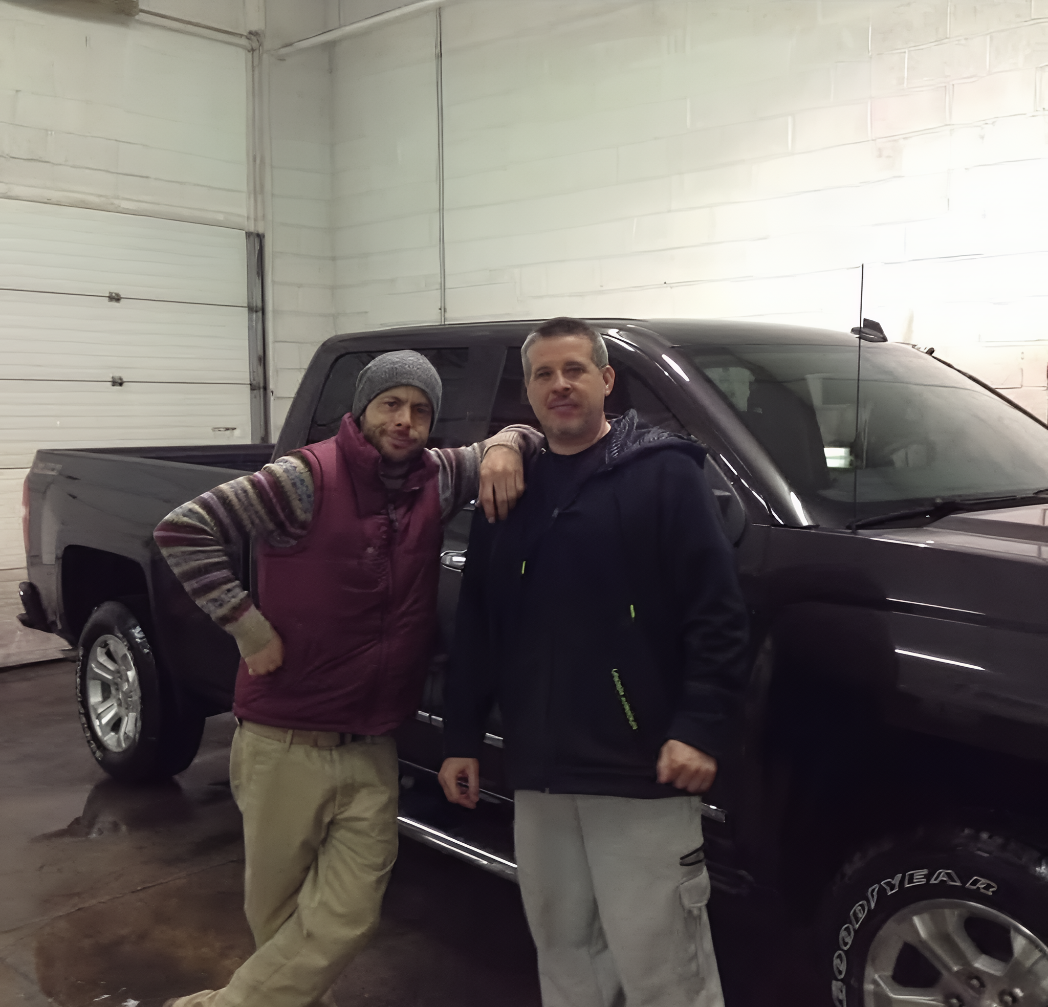 Two men standing next to a black pickup truck inside a garage, one with a beanie and striped sweater vest, the other in a black jacket.