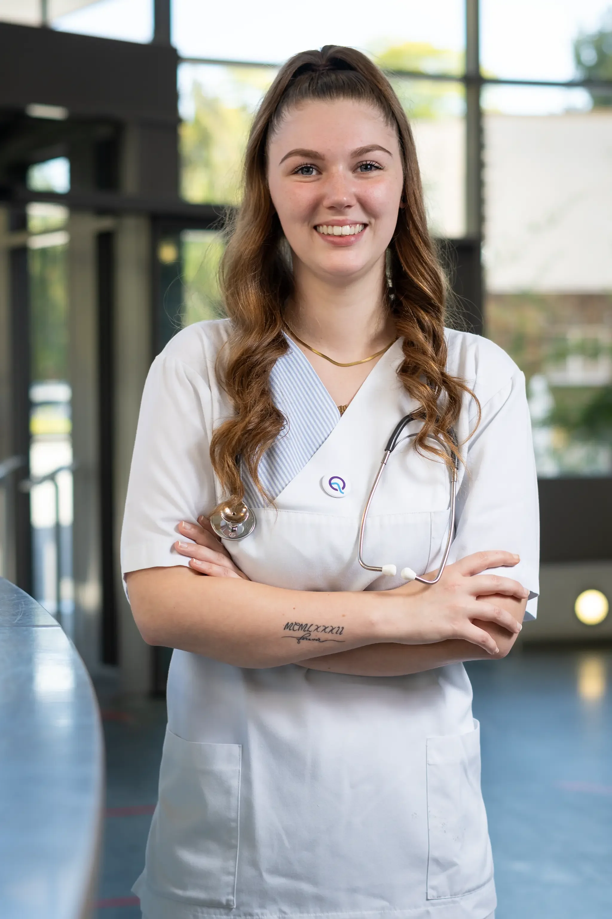 Eine junge Frau in einem weißen Arztkittel mit Stethoskop um den Hals steht mit verschränkten Armen in einem modernen Krankenzimmer und lächelt.