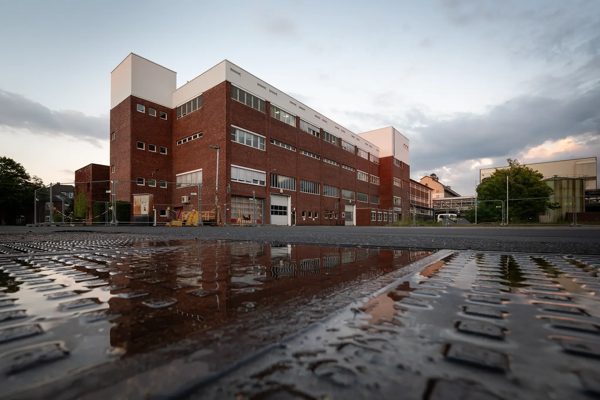 Ein mehrstöckiges, rotes Backsteingebäude auf dem Zanders-Gelände, im Vordergrund eine nasse, rifflige Metallebene mit einer Pfütze, die das Gebäude spiegelt, bei bewölktem Himmel.