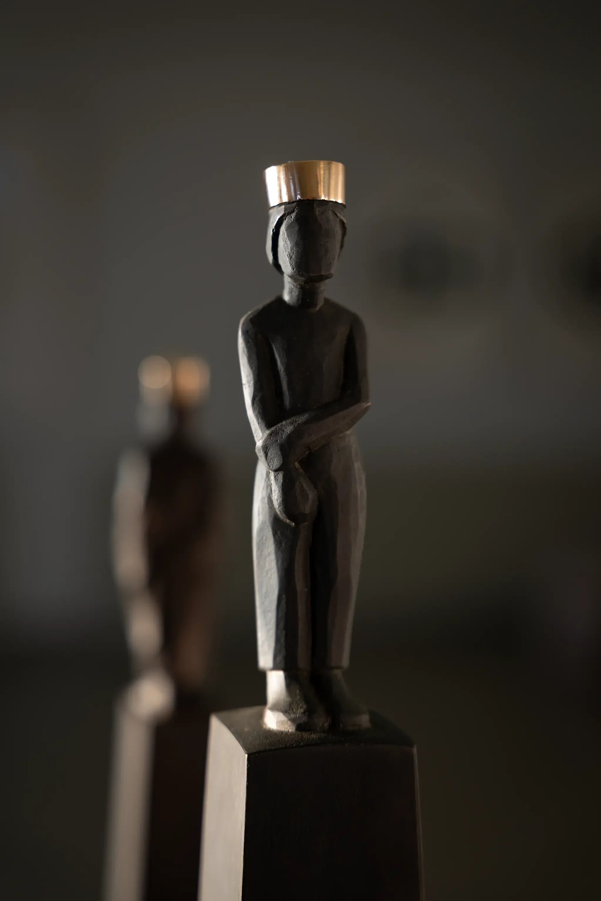 Skulptur eines männlichen menschlichen Körpers, der auf einem Sockel steht, mit einer goldfarbenen Krone auf dem Kopf, dunkle Holzmaserung, dunkler Hintergrund.