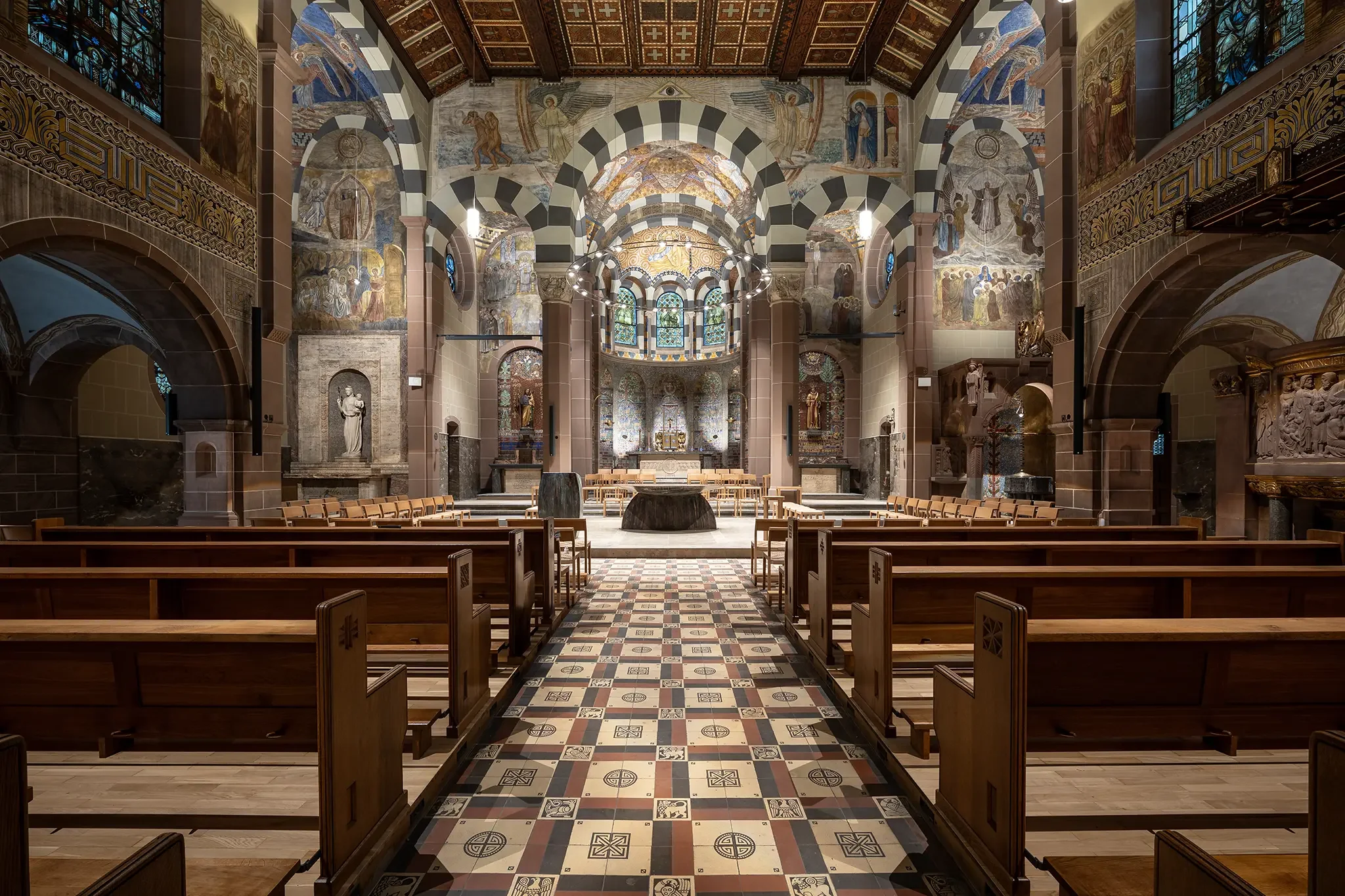 Innenansicht einer kunstvoll gestalteten Kirche mit gemustertem Boden, Holzbänken, bemalten Wänden und kunstvollen Fenstern im Hintergrund.