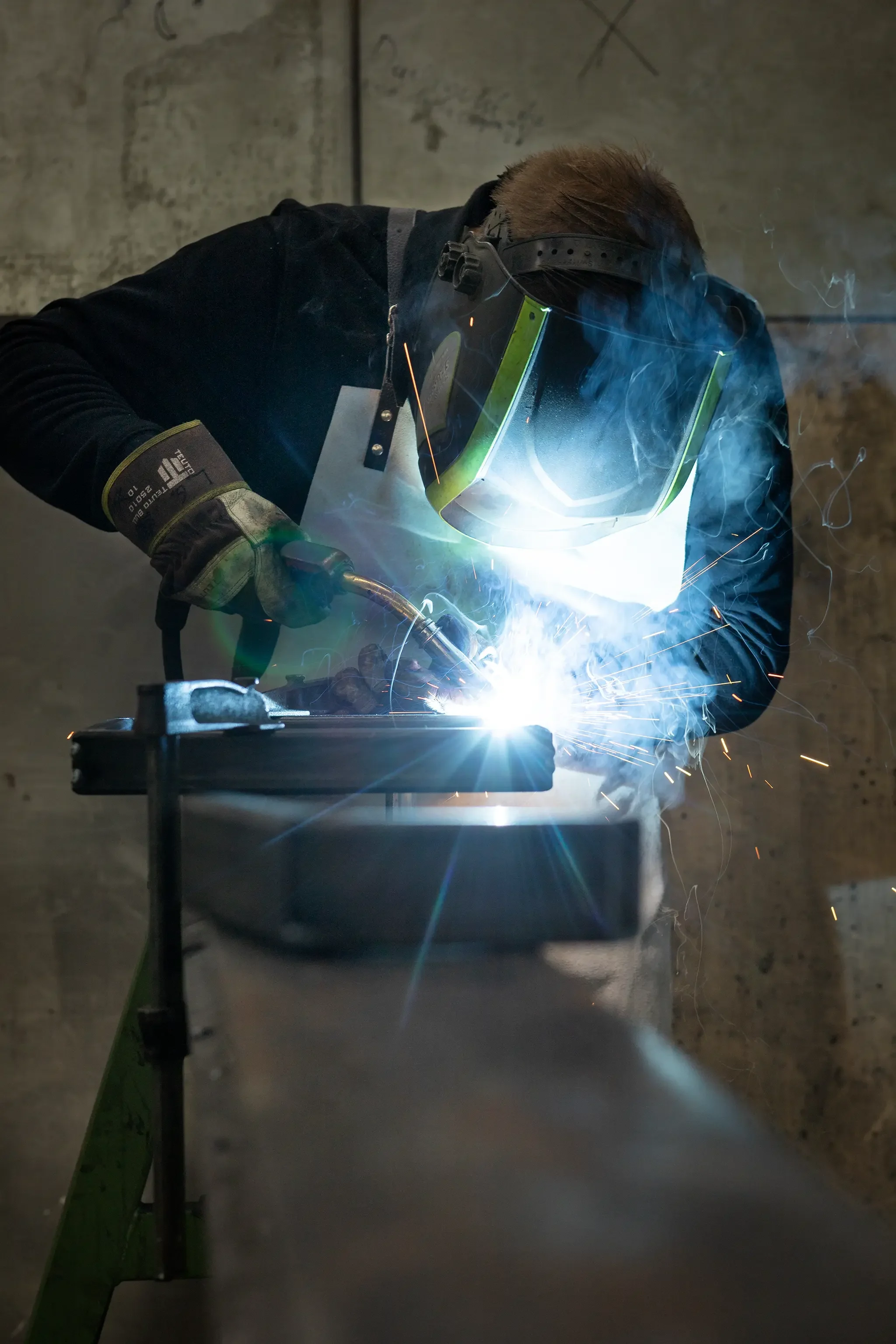 Schweißer in Schutzausrüstung beim Schweißen von Metall, mit blauem Licht und Funken, in einer Werkstatt.