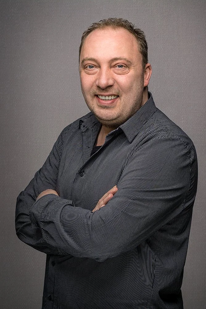 Porträt von Thomas Merkenich mit grauem Hemd, Arme verschränkt, vor grauem Hintergrund.
