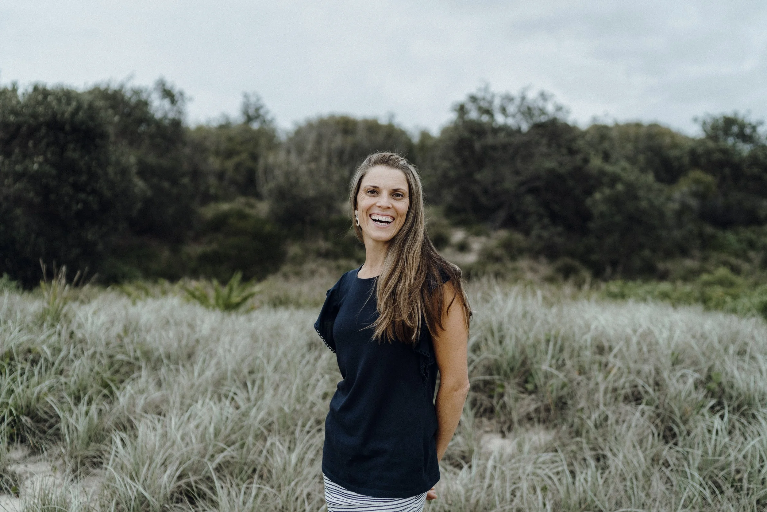 Melanie Woollam, Osteopath, standing outdoors in a grassy area, smiling at the camera, with trees and shrubs in the background.