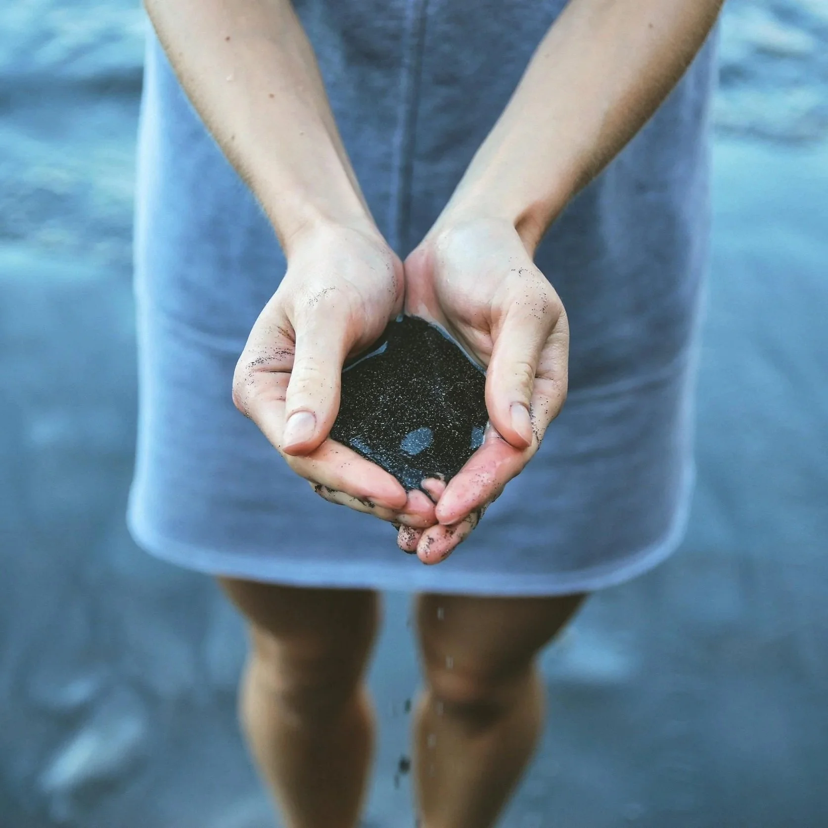 A person standing in shallow water holding a black rock with a small hole in the center, sand on their hands and around the rock.