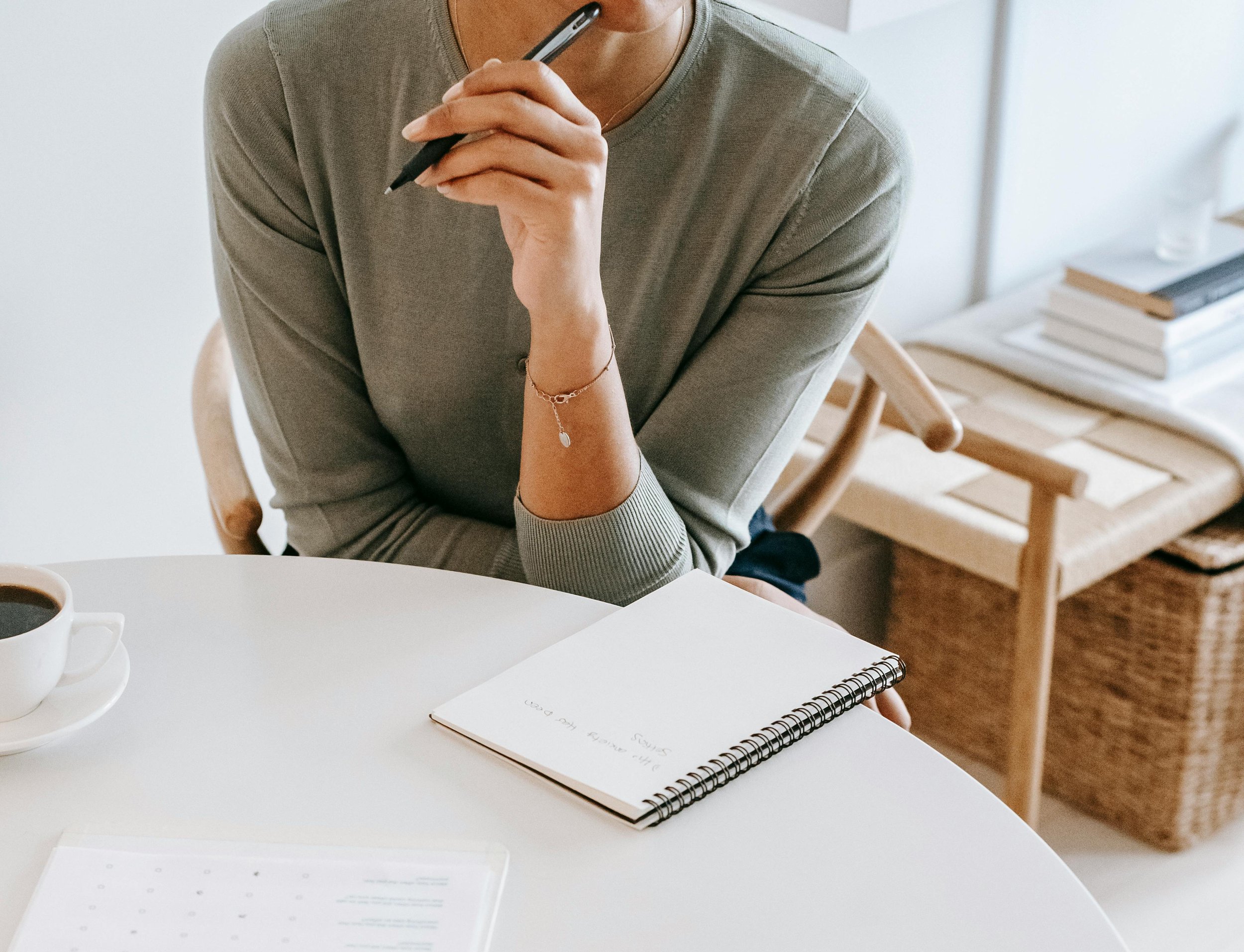 Person sitting at a white table, holding a black pen and writing in a spiral notebook, with a cup of coffee nearby and a stack of books and wicker basket in the background.