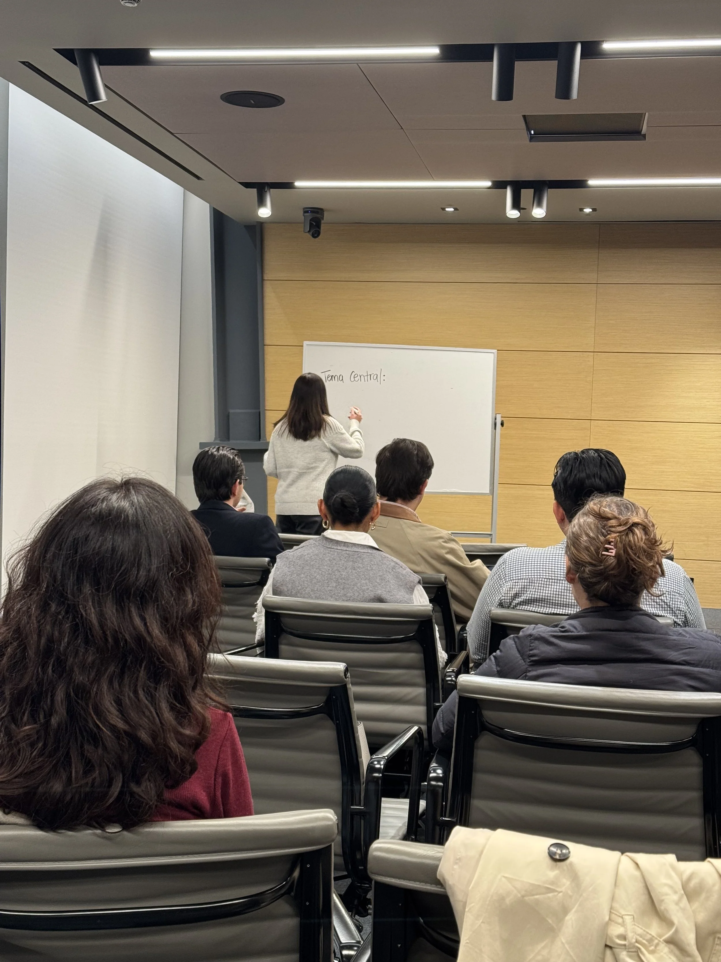 Clase en una sala de conferencia con varias personas sentadas observando a una instructora escribiendo en una pizarra blanca que dice 'Tema Central'.