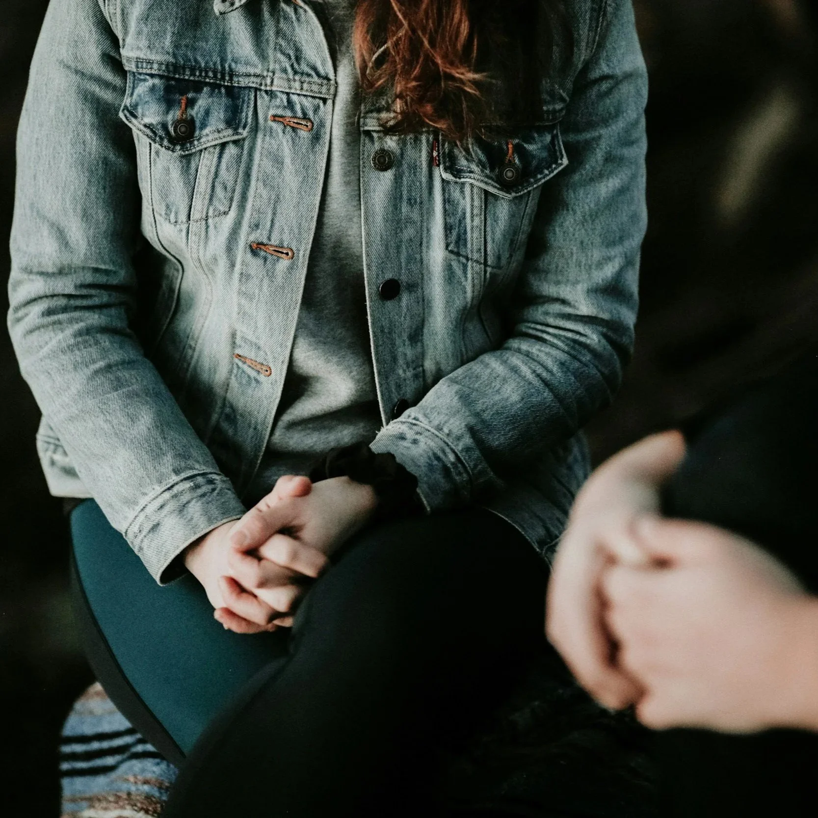A person wearing a light-colored denim jacket and gray shirt, with long brown hair, is sitting with hands clasped.