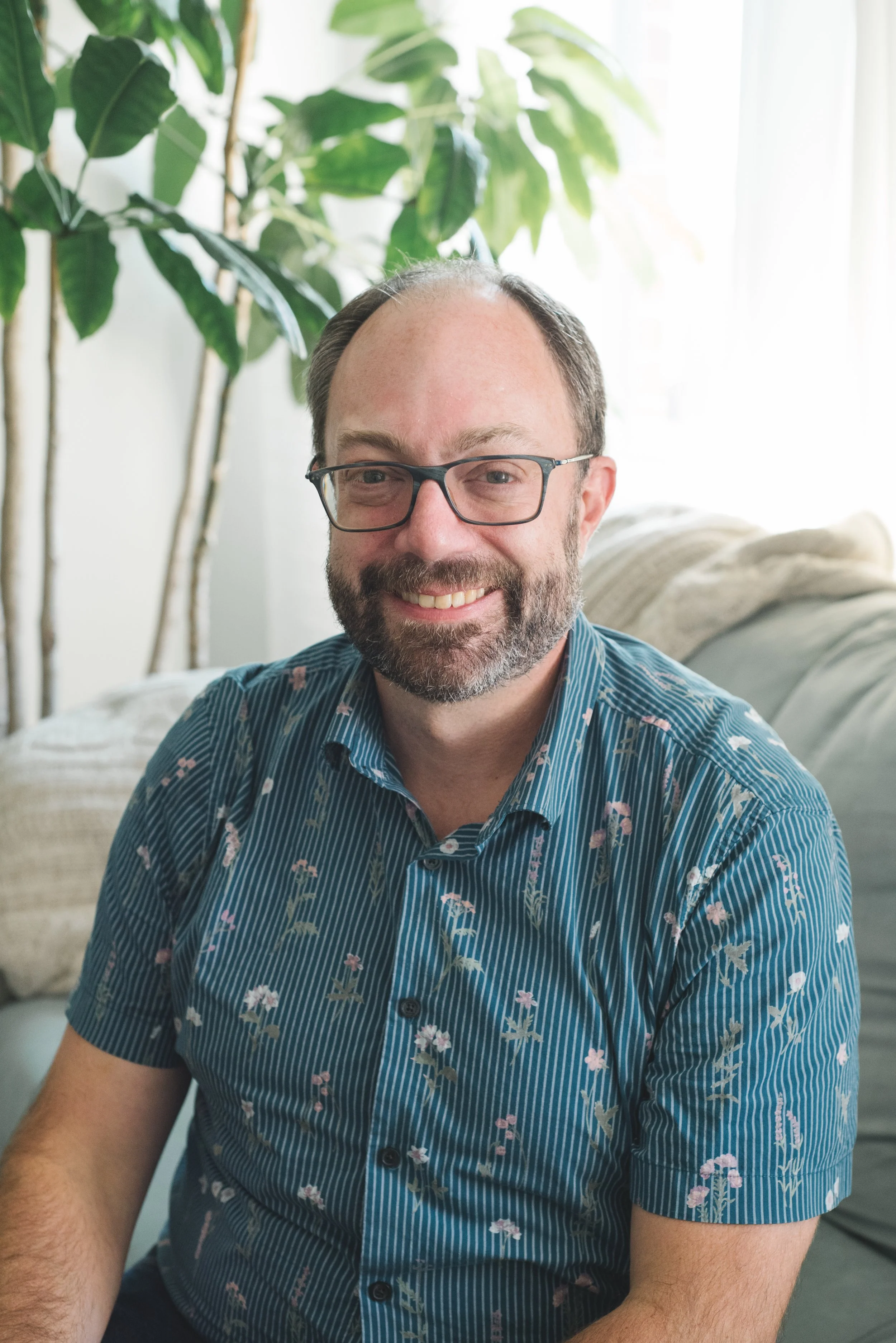 A man with glasses and a beard, smiling, wearing a black textured polo shirt with white trim, standing indoors in front of a decorative wall with multiple white window frames and some plants.