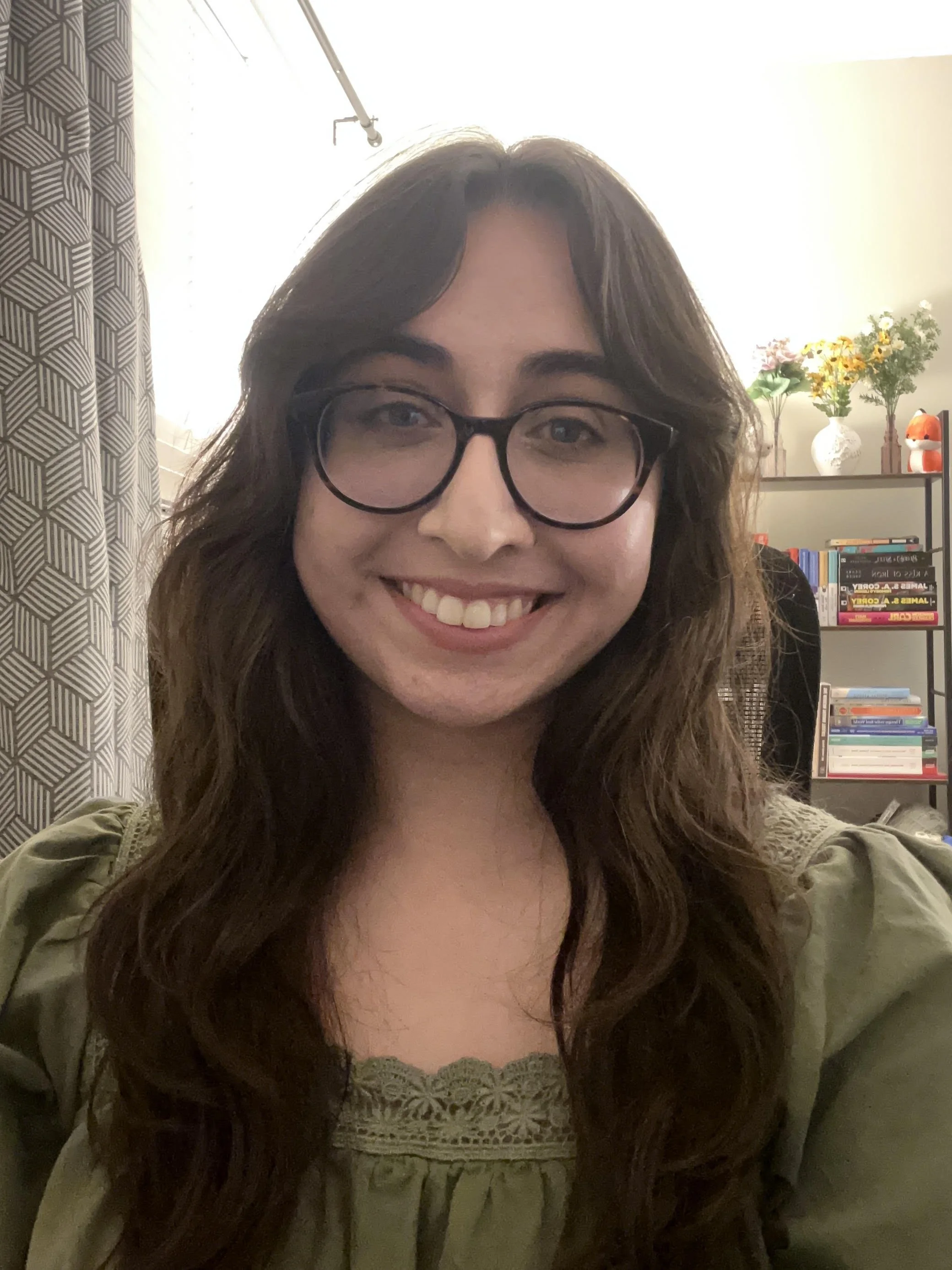 A woman with long brown hair and glasses smiling indoors, with a bookshelf and flowers in the background.