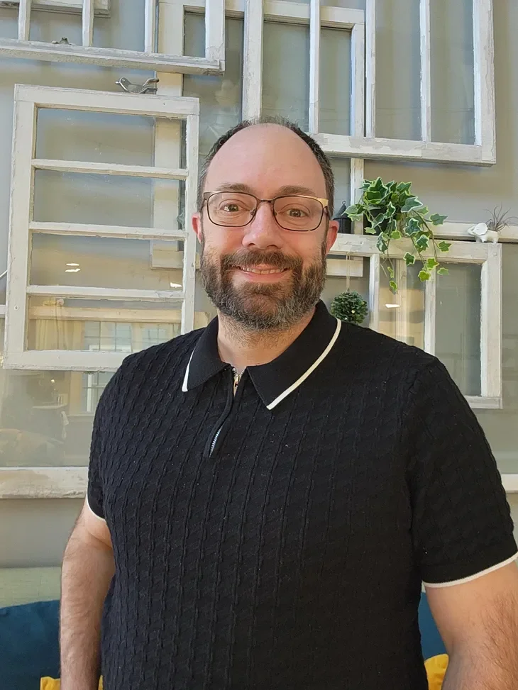 A man with glasses and a beard smiling, wearing a black polo shirt with white trim, standing indoors in front of a wall with decorative window frames and greenery.