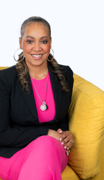 A smiling woman sitting on a yellow couch, wearing a black blazer, bright pink dress, and large hoop earrings.