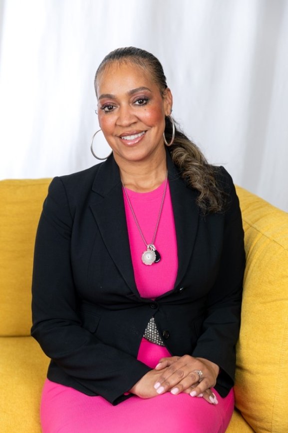 Woman sitting on a yellow couch, smiling, wearing a black blazer over a pink dress, hoop earrings, and necklaces, with a white curtain background.