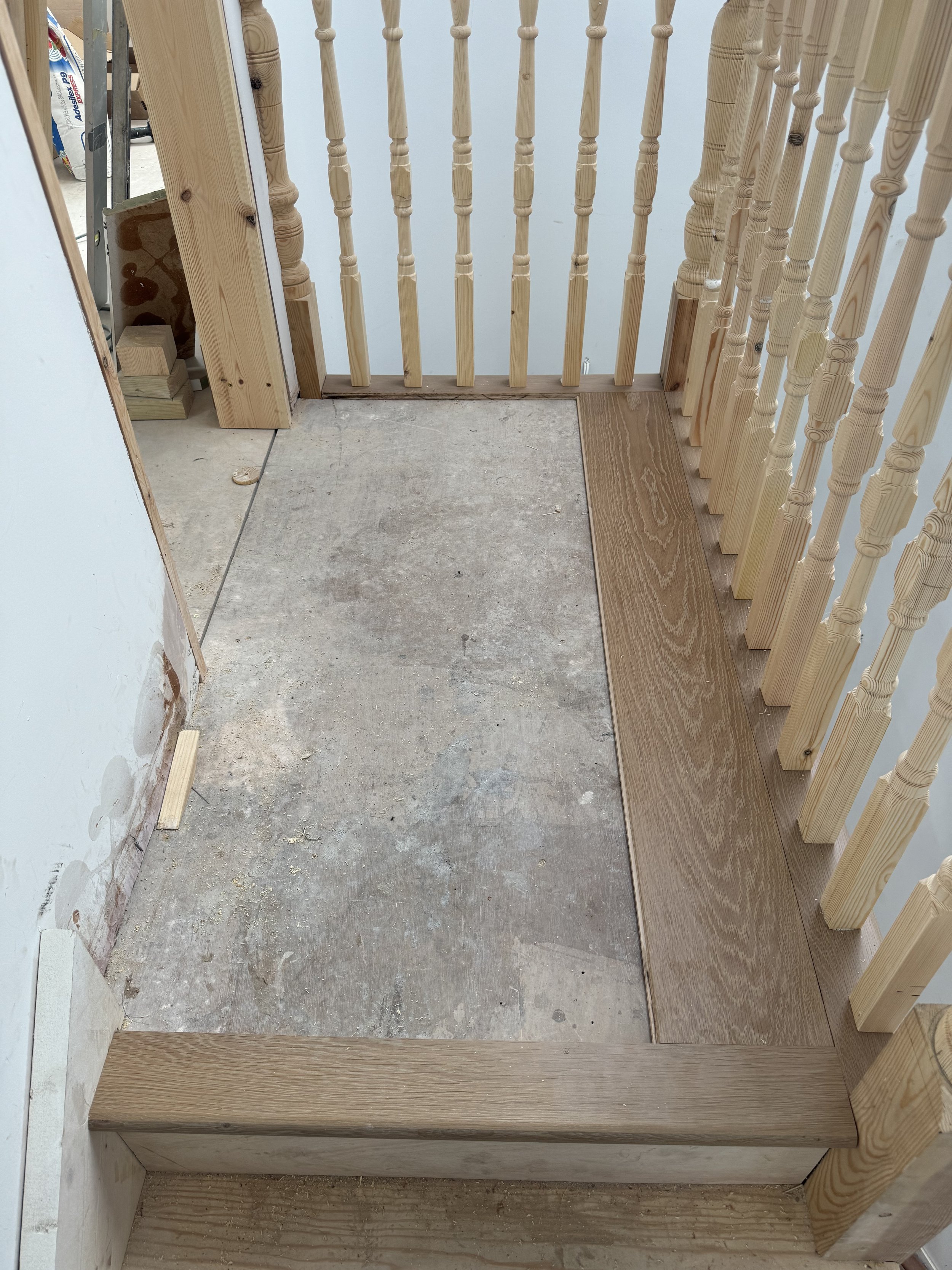 Close-up of a staircase with wooden railing, partially finished floor with plywood, and construction materials in the background.