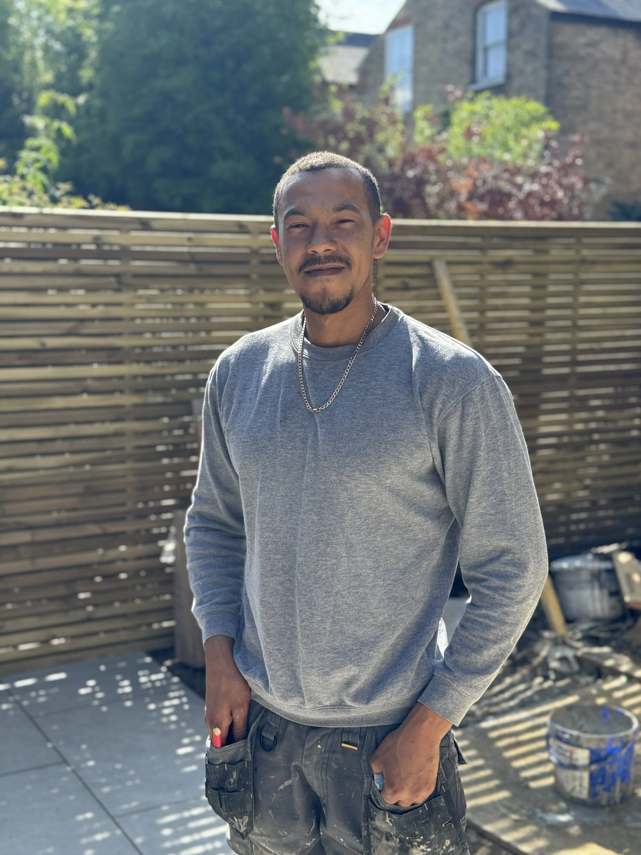 A man standing outdoors wearing a gray long-sleeve shirt and camouflage pants, smiling slightly, with a wooden fence, trees, and a house in the background.