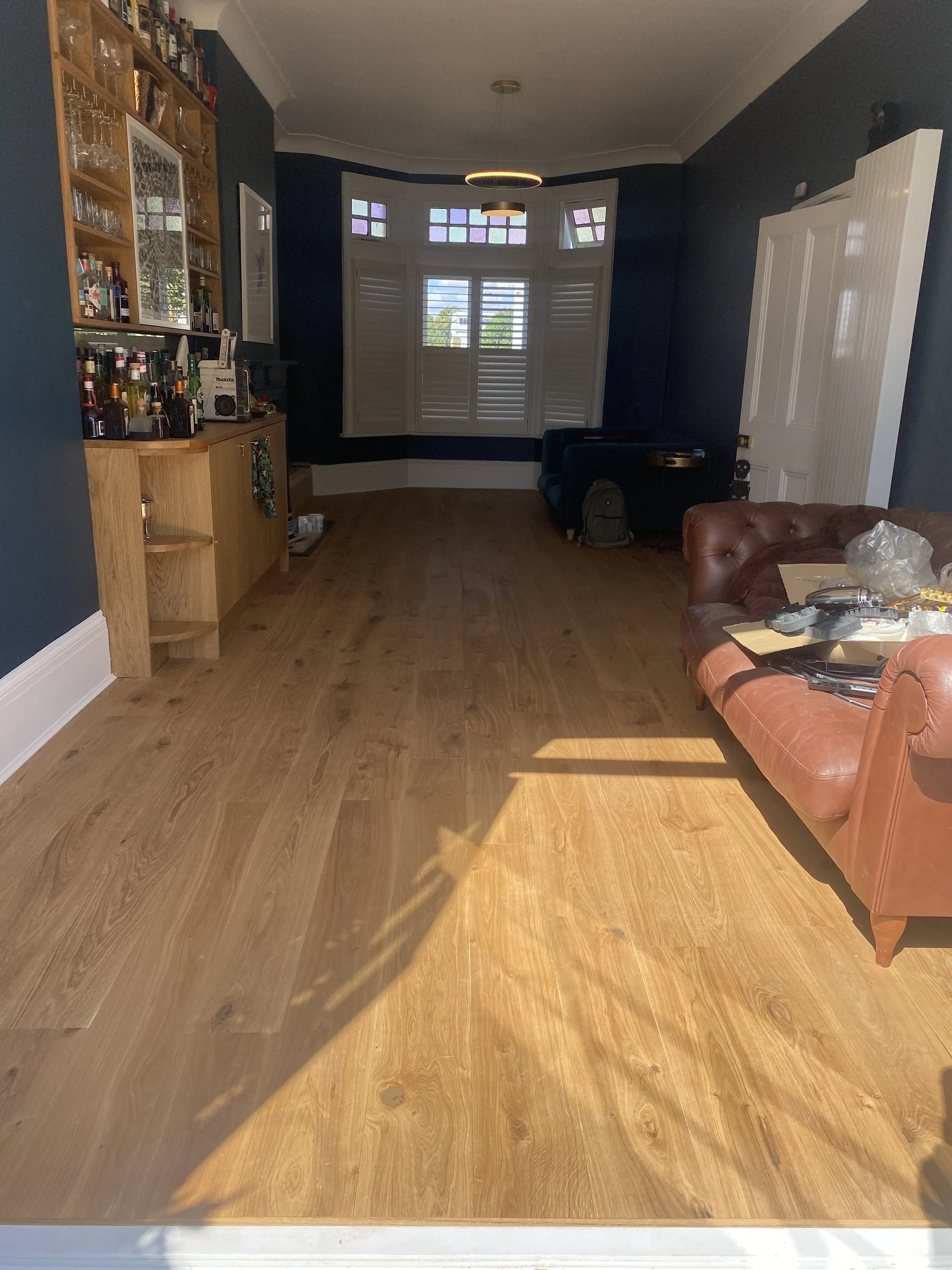 Interior of living room with wooden floor, dark blue walls, a large bay window with shutters, a brown leather sofa, a bookcase with bottles and glassware, and an assortment of items on the sofa.