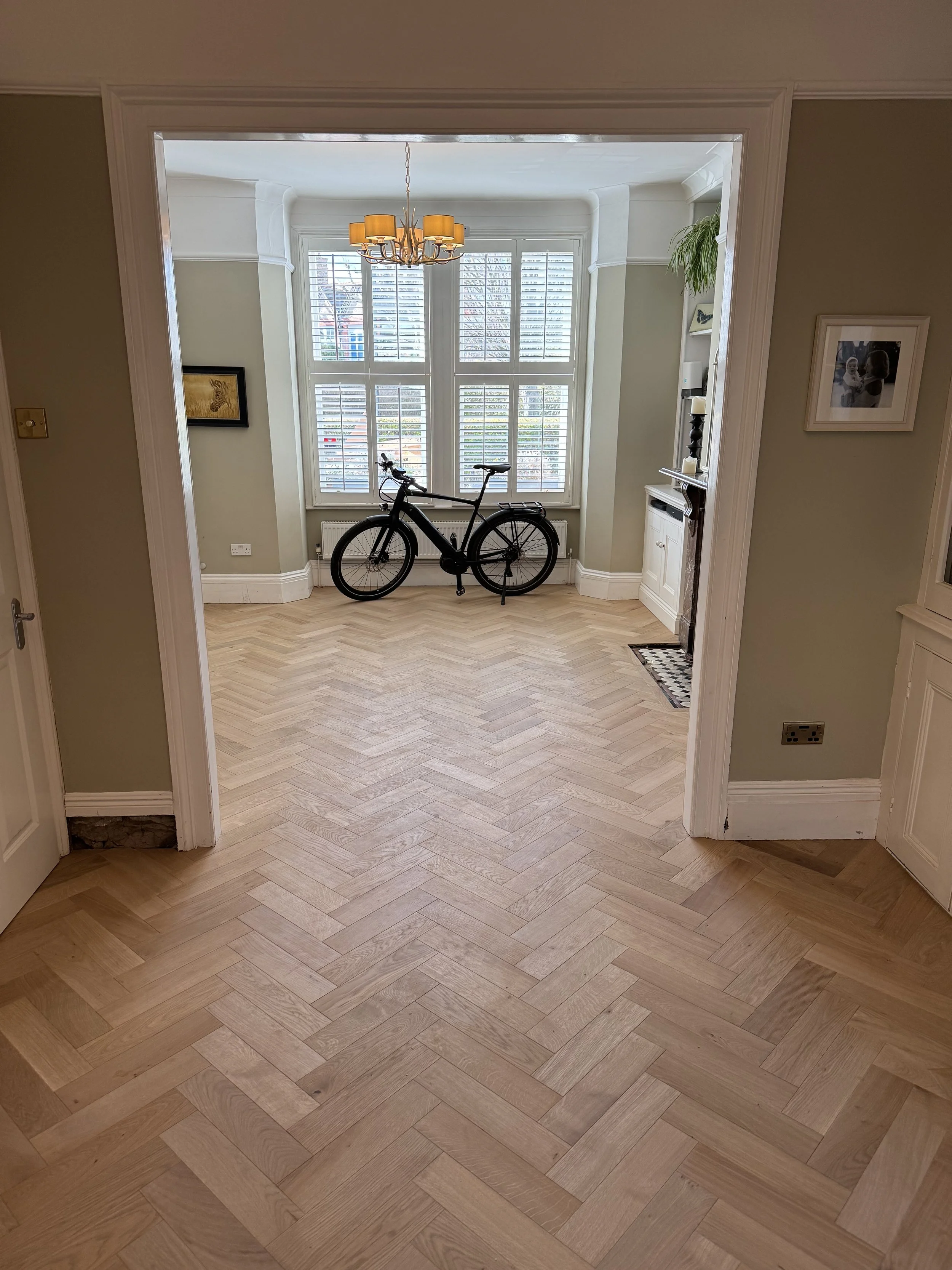 View through a doorway into a room with a bicycle leaning against a bay window with white plantation shutters. The room has a chandelier, a potted plant, and built-in shelves.