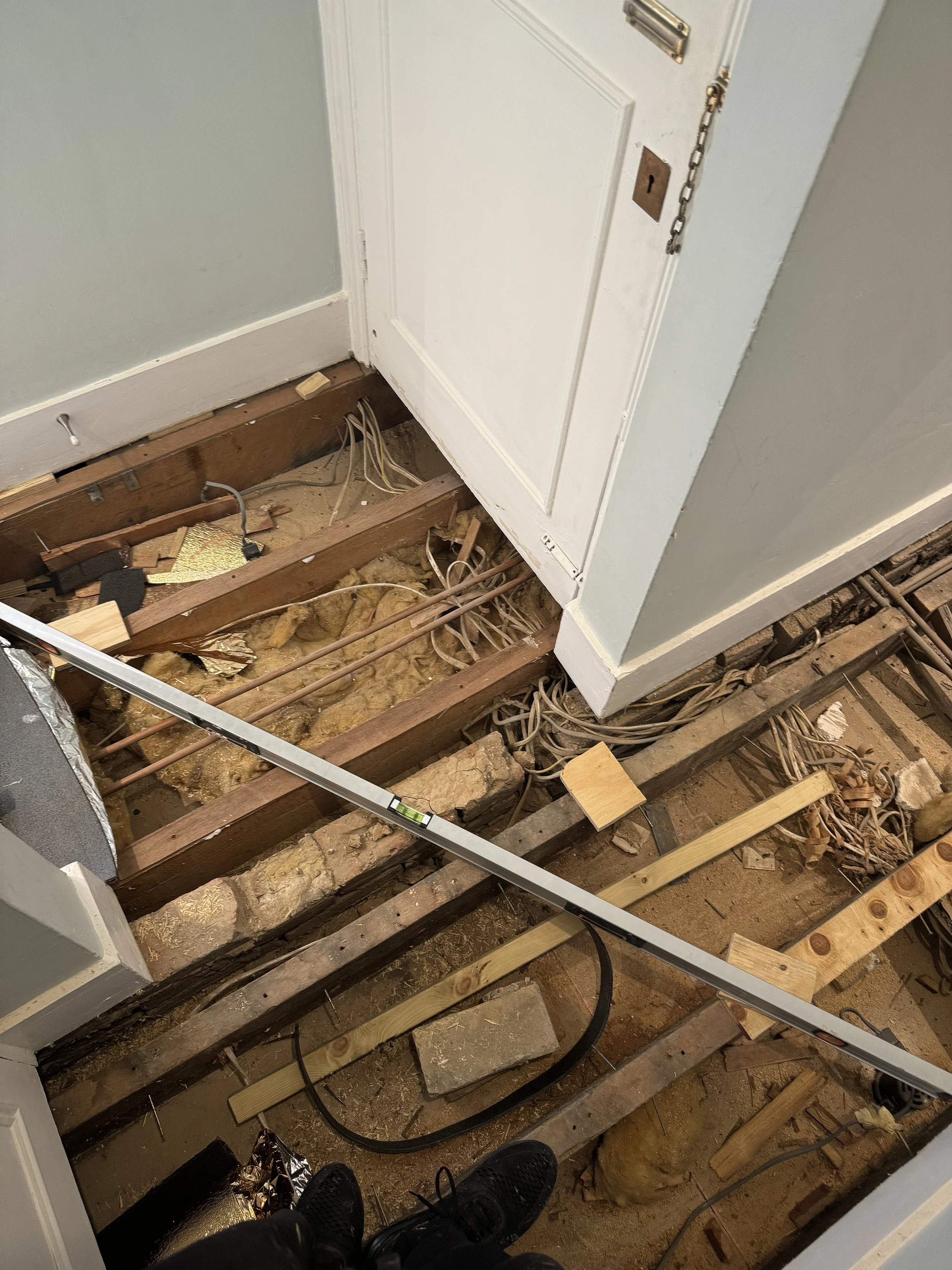View of a room with the flooring removed, exposing wooden beams, electrical wiring, insulation, and construction tools.