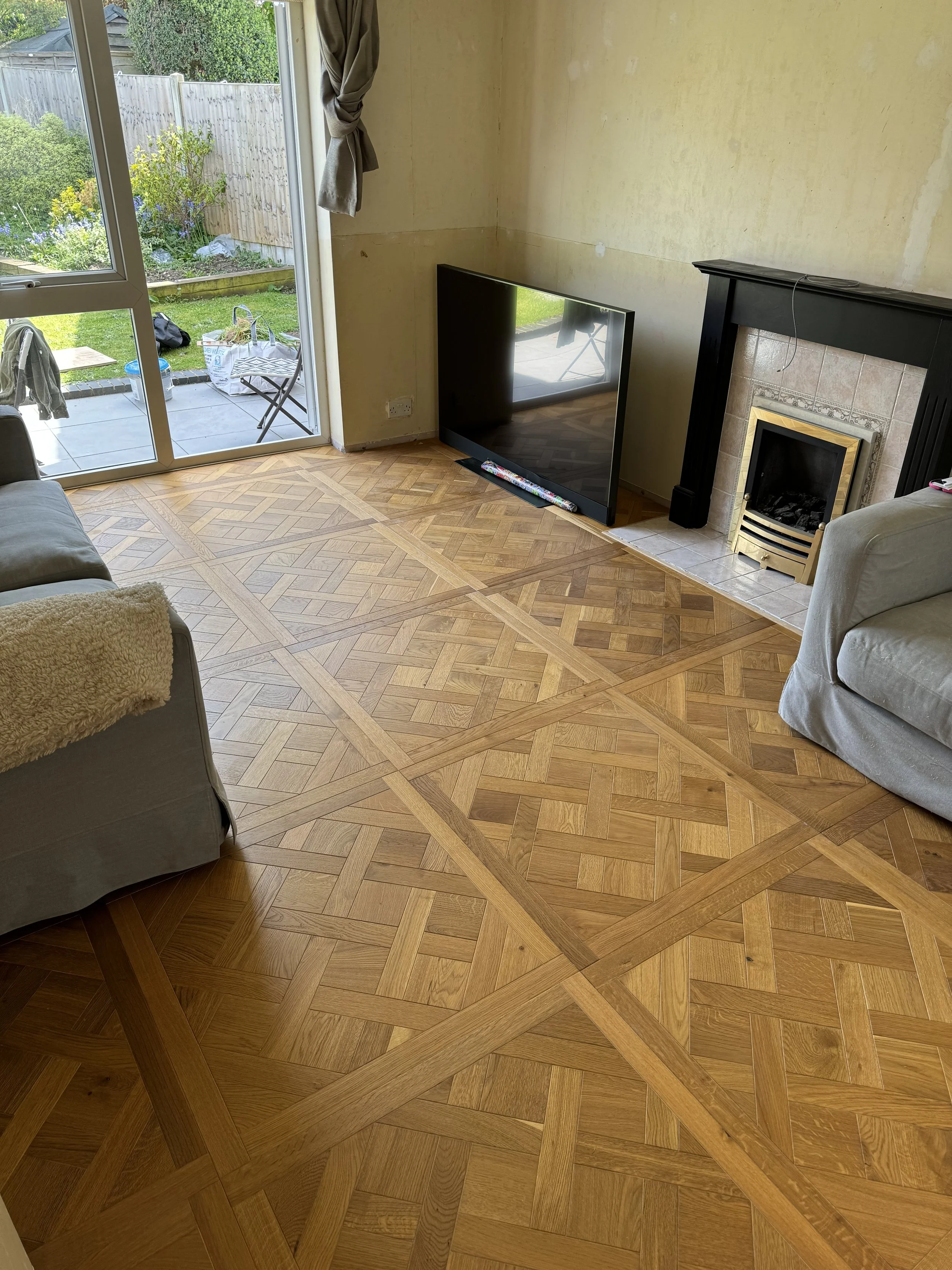 Living room with parquet flooring, a fireplace with black mantel, a flat-screen TV on the floor near the fireplace, and sliding glass doors leading to a garden with outdoor chairs and a table.