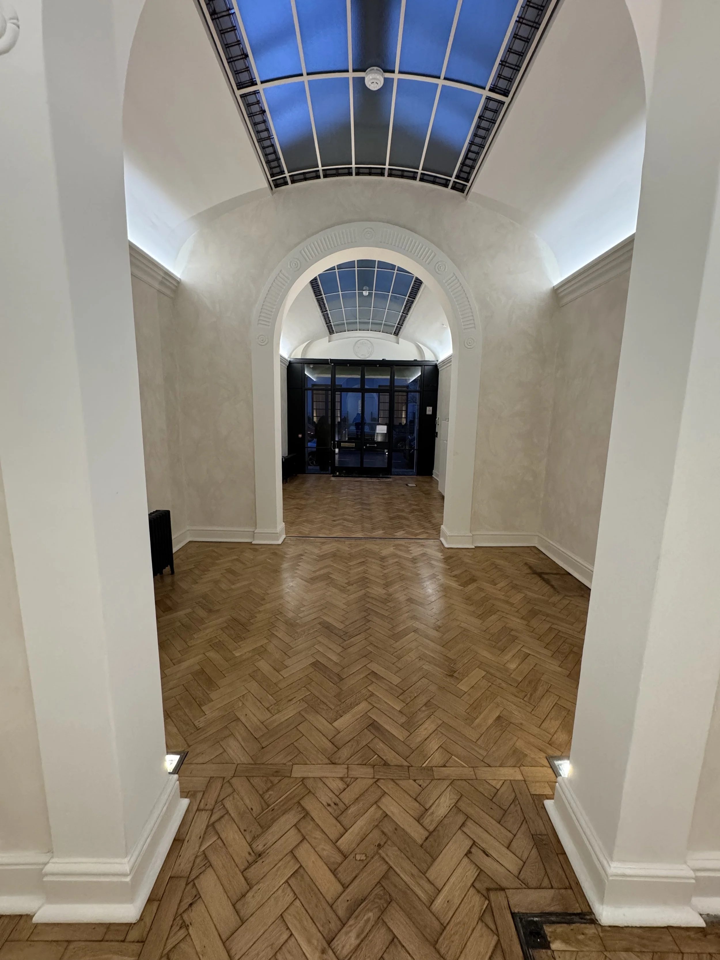 An indoor hallway with wooden herringbone floor, white walls, arched doorways, and a ceiling with skylights and a fire alarm. The corridor leads to a glass door at the end, showing an outside area.