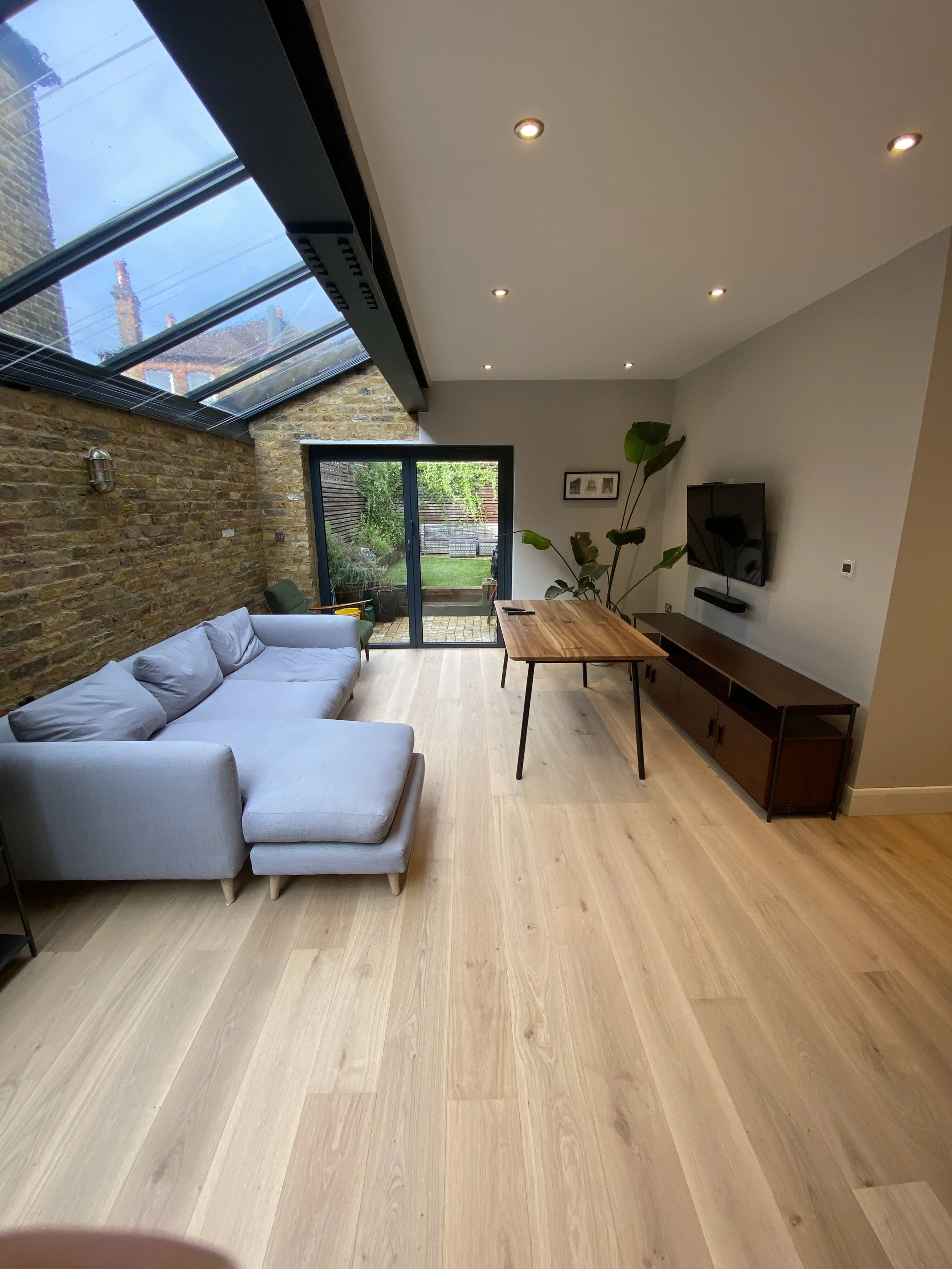 Living room with a gray sofa, wooden flooring, brick wall on the left, sliding glass door opening to a garden, and a wall-mounted TV with a wooden media console.