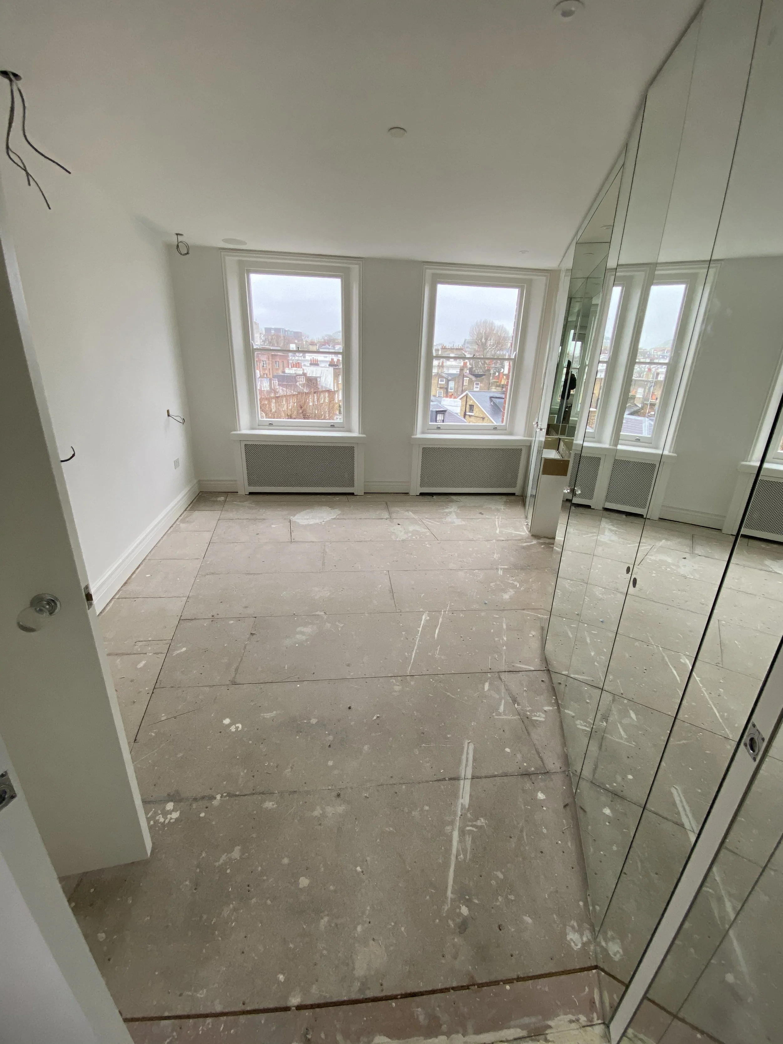 Empty room with three large windows overlooking a cityscape, white walls, mirrored closet doors, and a bare concrete floor with paint splatters.