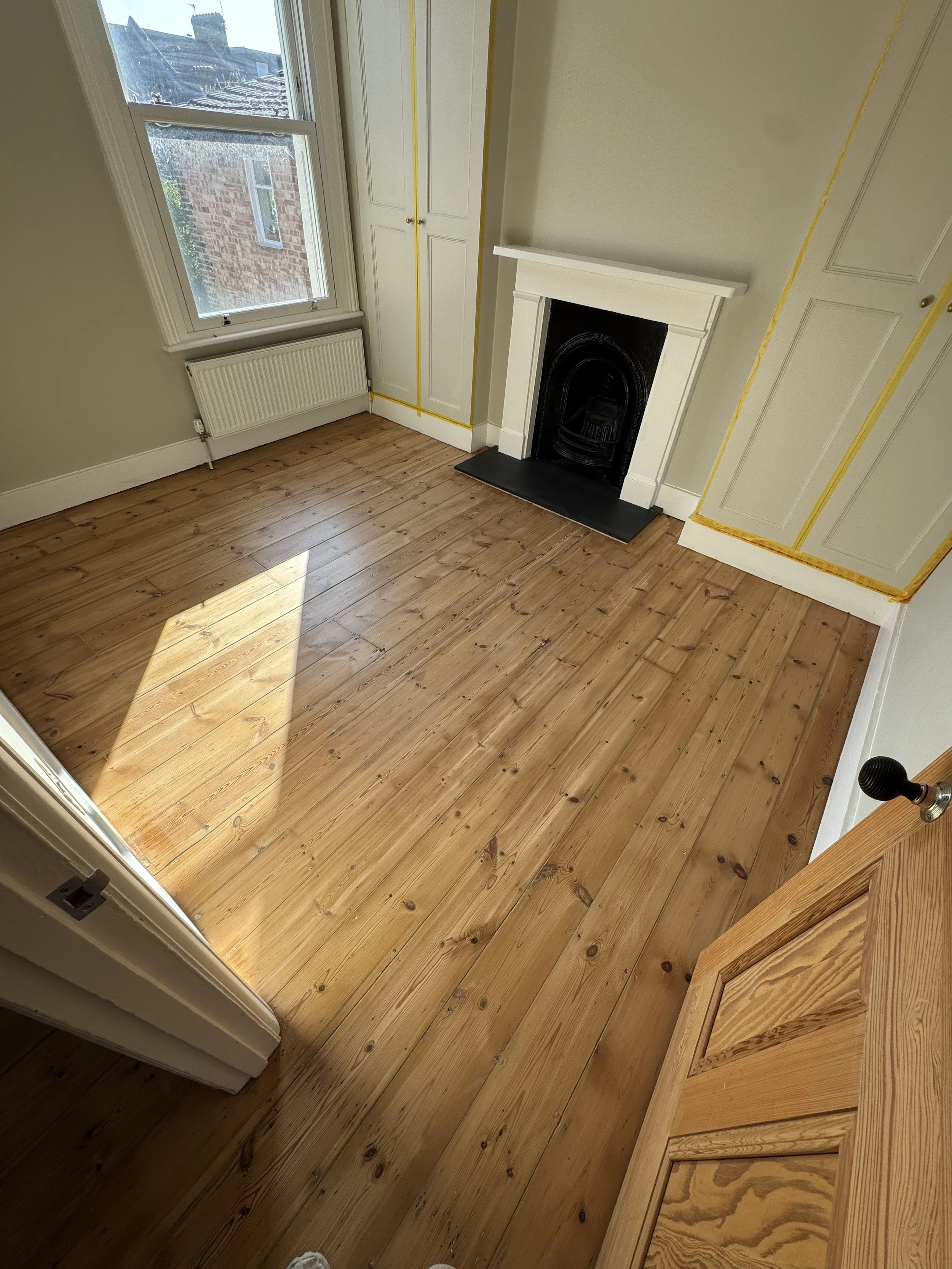 Empty room with wooden floor, white walls, a black fireplace, a window letting in sunlight, a white radiator, and built-in cabinets with yellow tape outlining the edges.