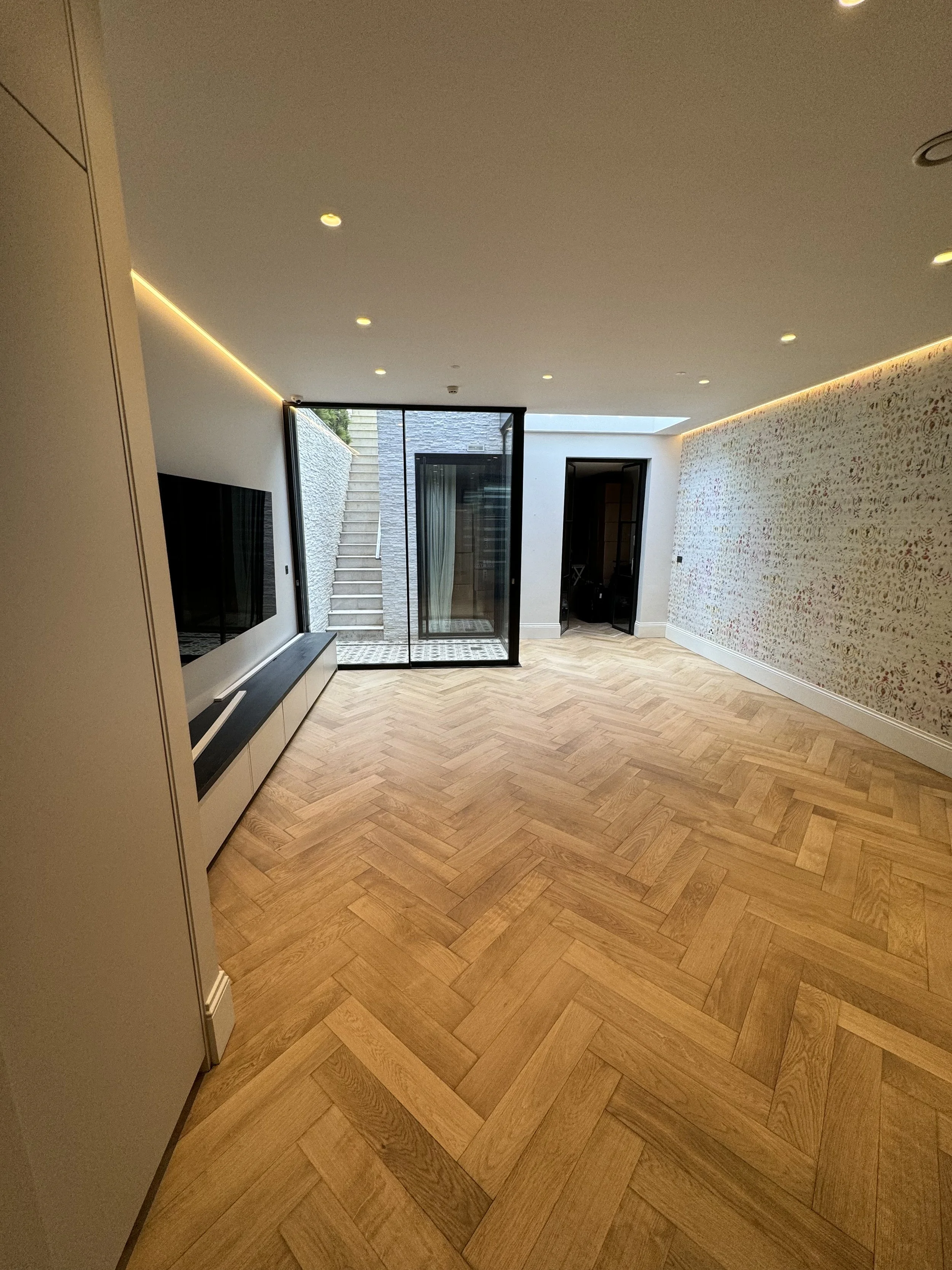 Modern living room with wooden herringbone flooring, a flat-screen TV, and large glass doors leading outside. The room has recessed ceiling lights and a curved, decorative wall.