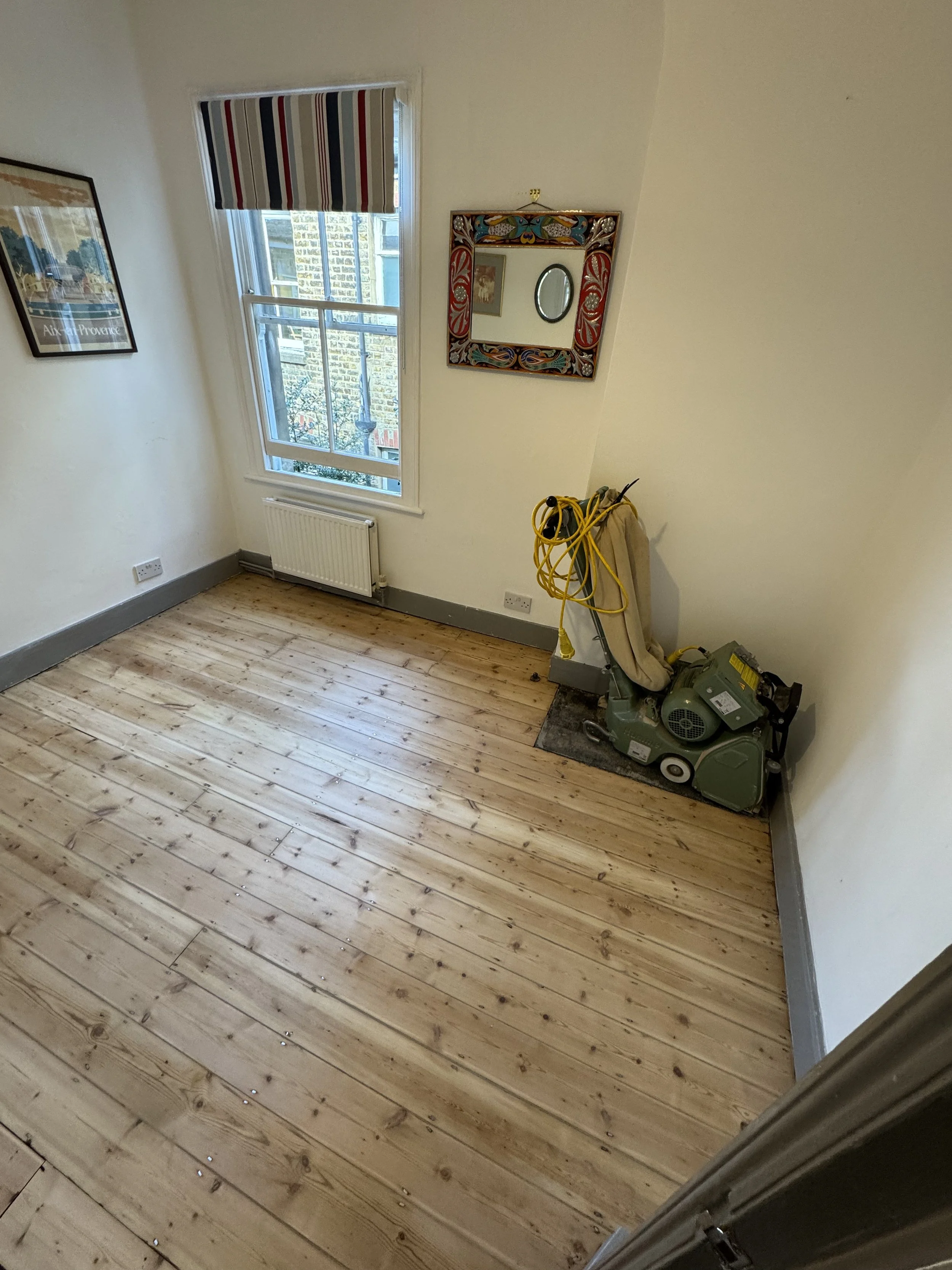 An empty room with wooden floorboards, a window with a striped curtain, a radiator, a colorful framed mirror, a wall painting, and a floor sander machine in the corner.
