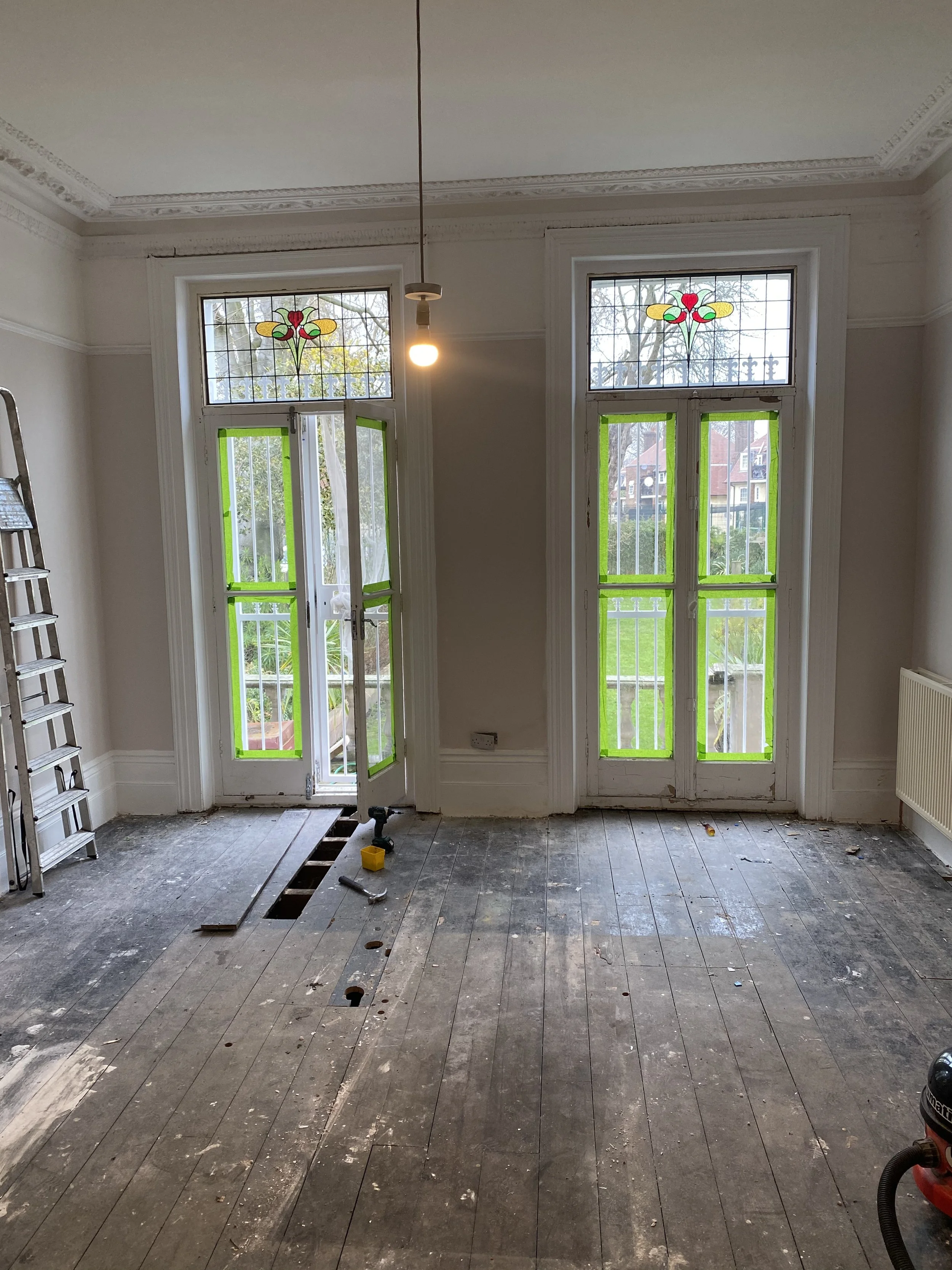 Room under renovation with unfinished wooden floor, a ladder on the left side, a hanging light bulb from the ceiling, and two large windows with stained glass and green tape on the window frames, showing a green yard outside.