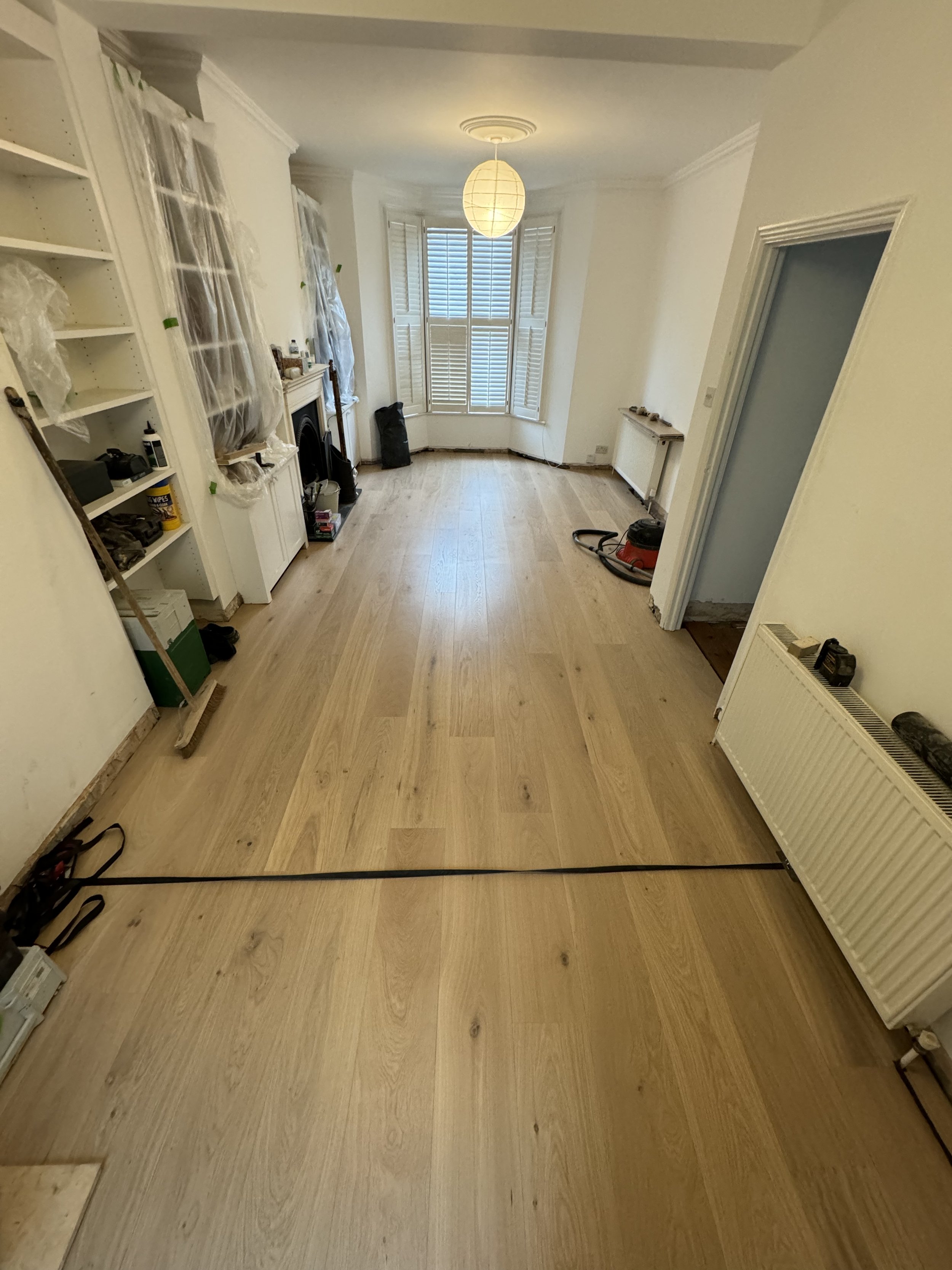 Living room with unfinished flooring and construction tools, a large bay window with white shutters, and a white ceiling with a paper lantern light fixture.