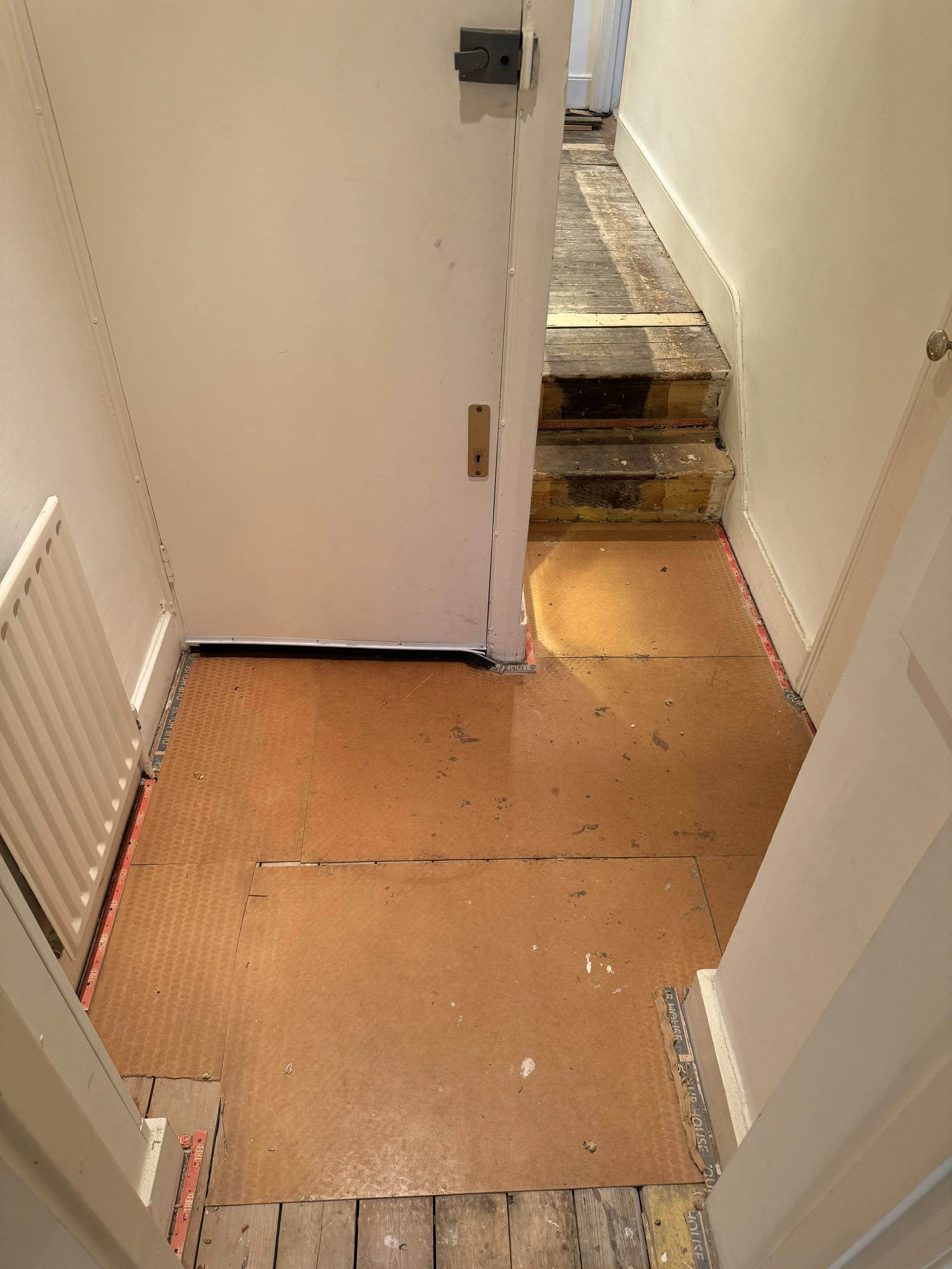 An interior area with subflooring and stairs, entrance to a room with a white door, an orange-colored underlayment, and wooden steps at the top of the stairs.
