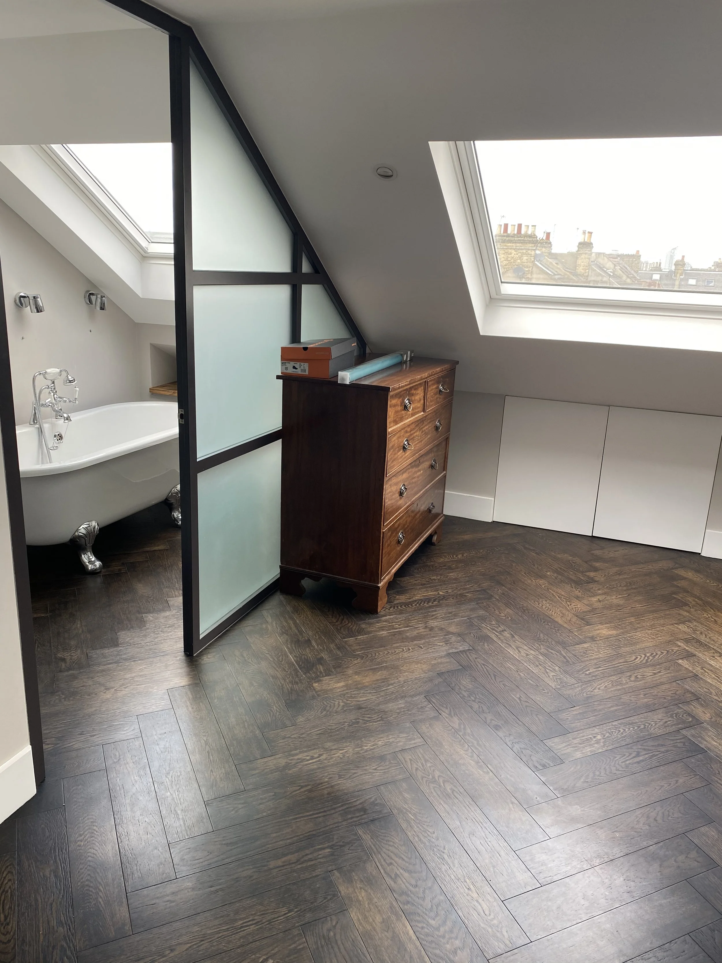 An attic room with dark wood flooring, a sloped ceiling with skylights, a wooden dresser, a partially visible bathtub separated by frosted glass panels, and white cabinets.