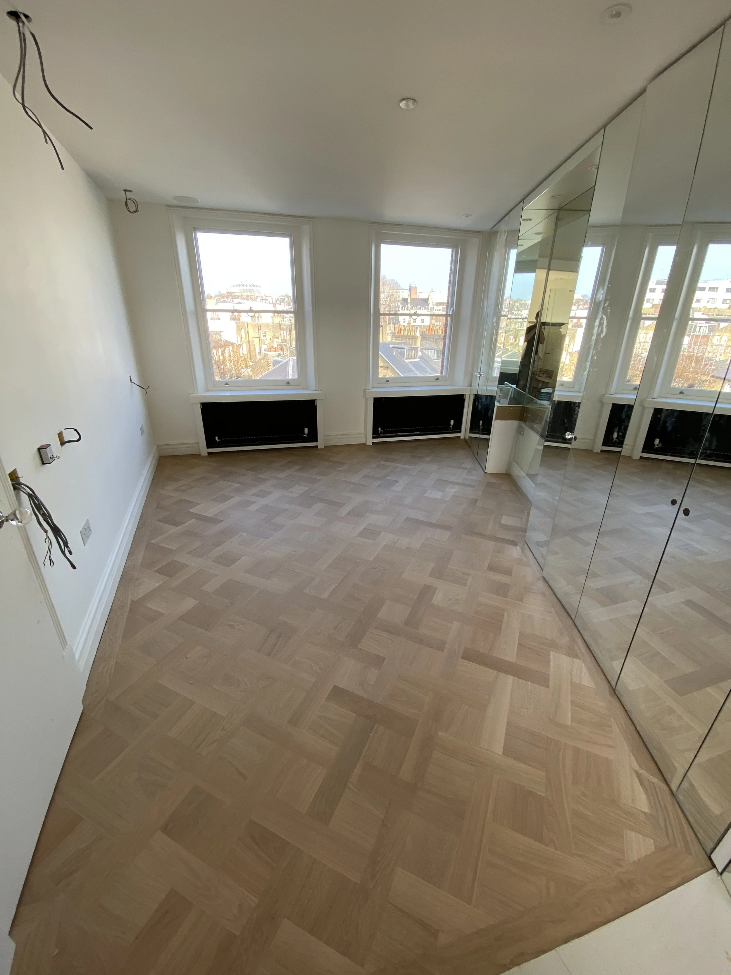 Empty room with large windows, parquet wood flooring, and mirrored closet doors, with some wiring hanging from the ceiling.