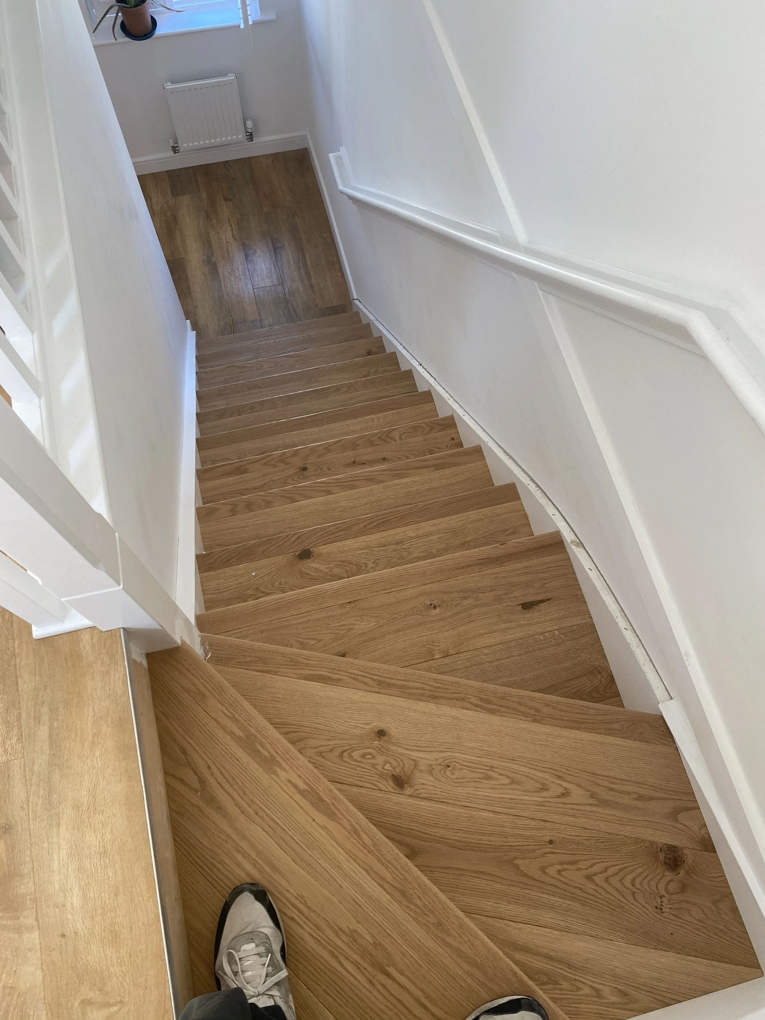 View looking down a wooden staircase from the top, with a person's sneakers visible at the top edge, white walls, a white handrail on the right, a small window at the bottom, and a wooden floor at the bottom of the stairs.